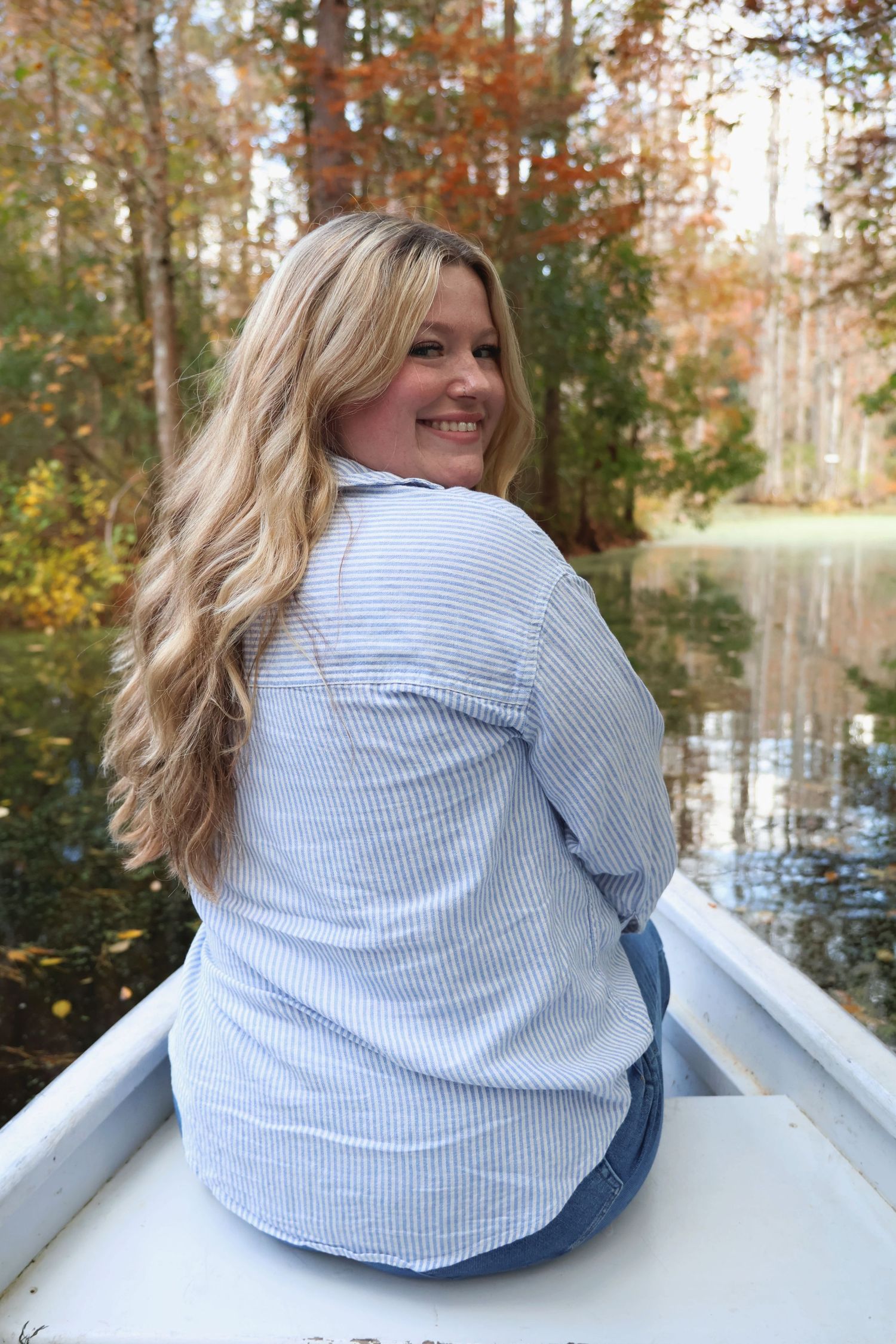 A woman with long blonde hair sitting in a small boat on the water, looking back over her shoulder with autumn trees in the background.