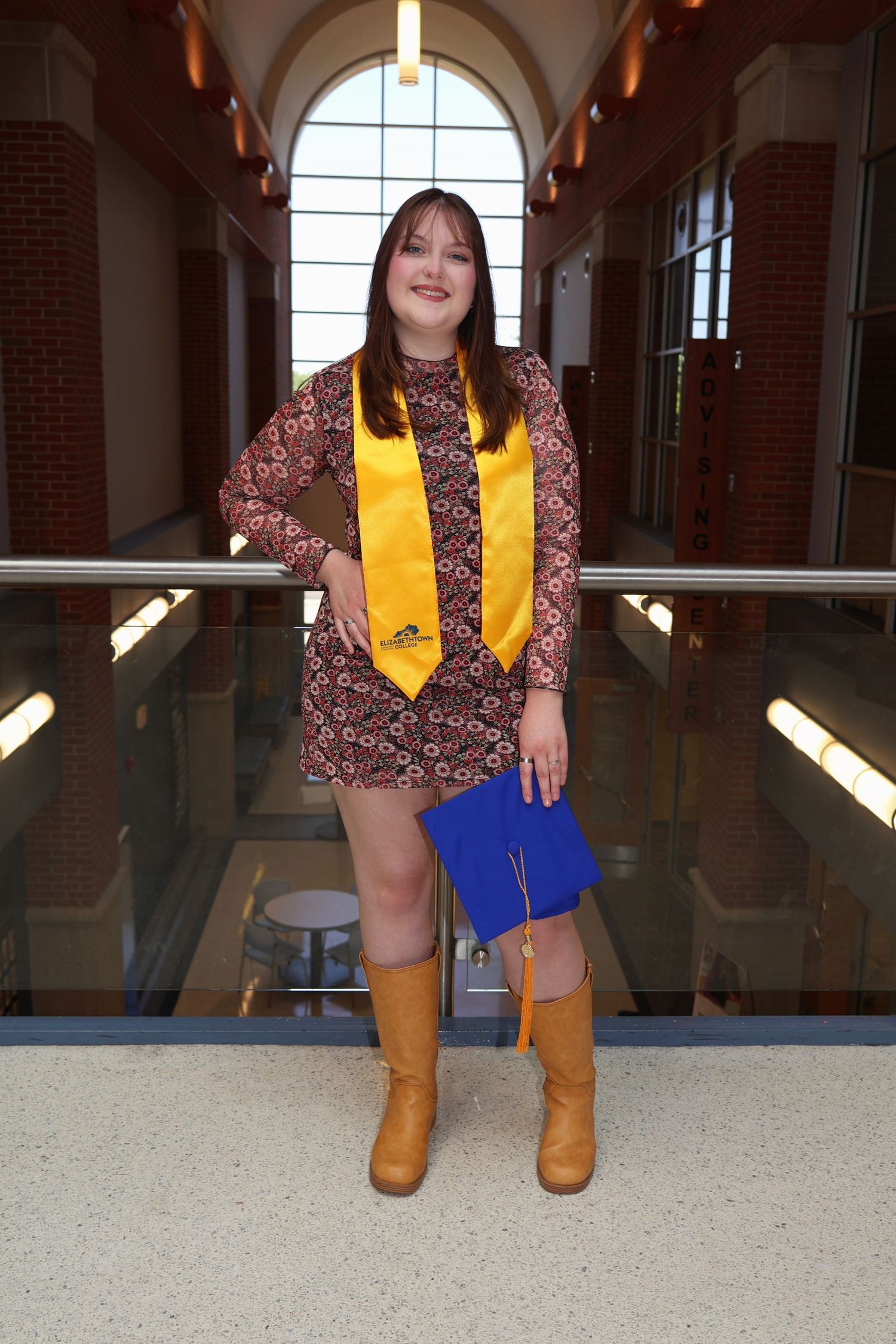 Full-length portrait of a graduating woman standing on an indoor walkway, wearing a yellow stole and holding her blue graduation cap.
