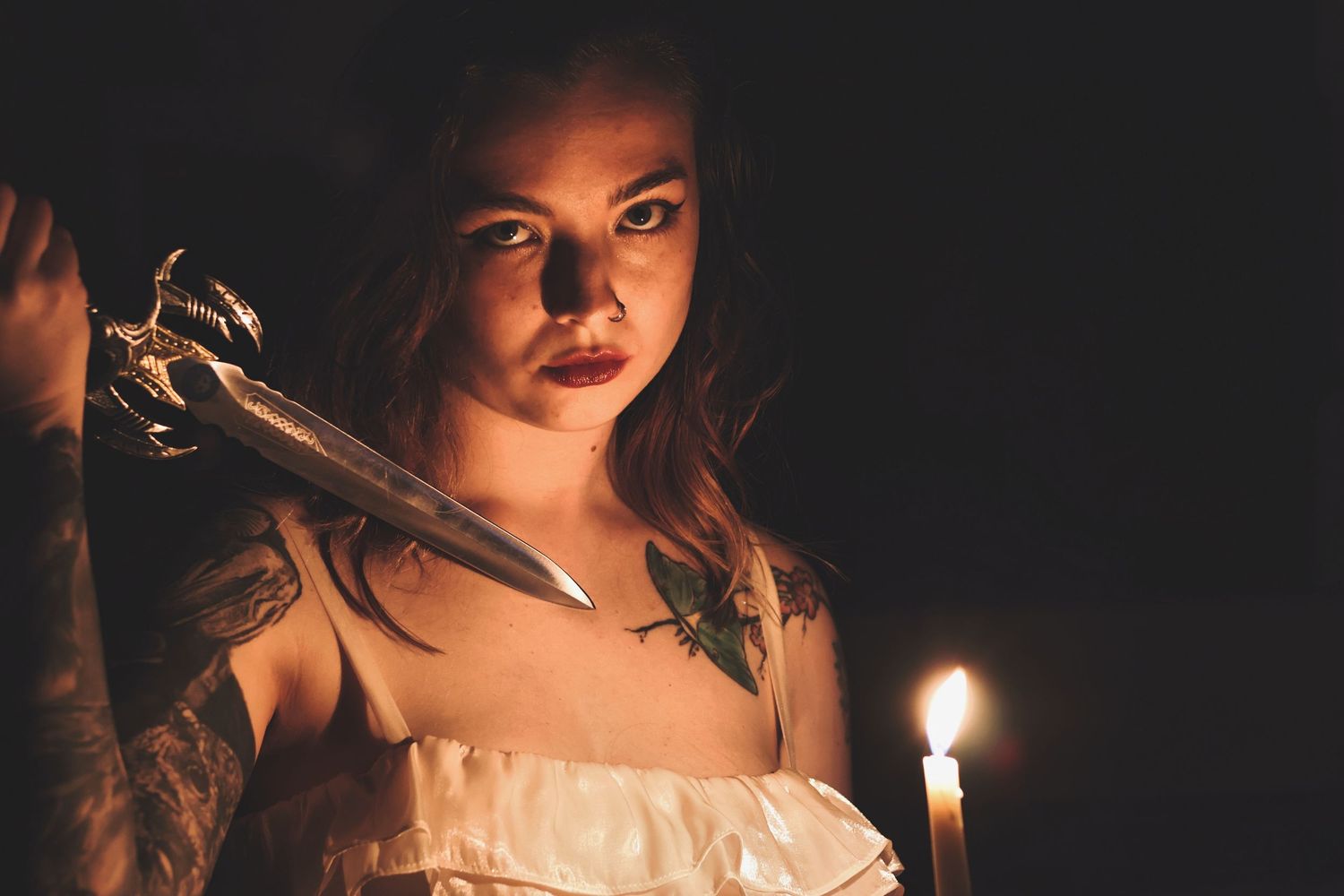 In a darkly lit room, a woman with a fierce expression brandishes an ornate dagger near her shoulder, illuminated by a single burning candle