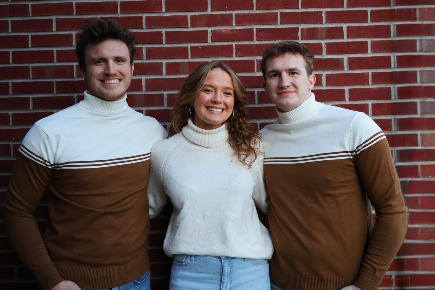 Three siblings posing together in matching color-coordinated turtlenecks in front of a red brick wall.
