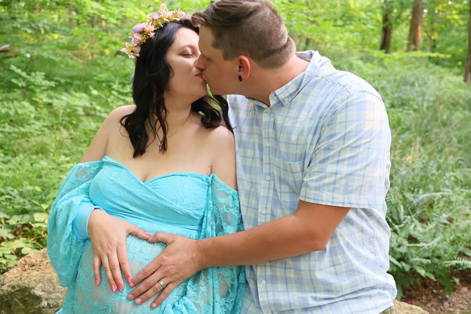 A man kissing a pregnant woman in a flower crown and cyan off-the-shoulder dress as they both hold her belly outdoors.