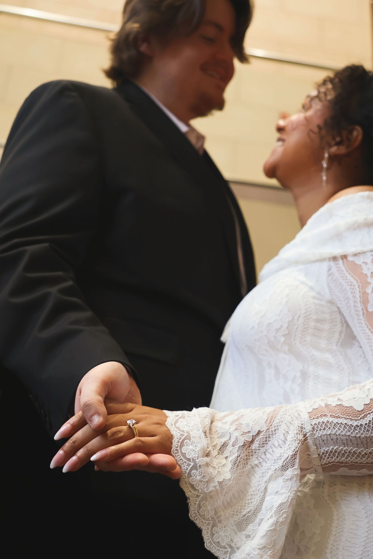 Low-angle shot of a newlywed couple holding hands and looking lovingly at each other, showcasing the bride's wedding ring.