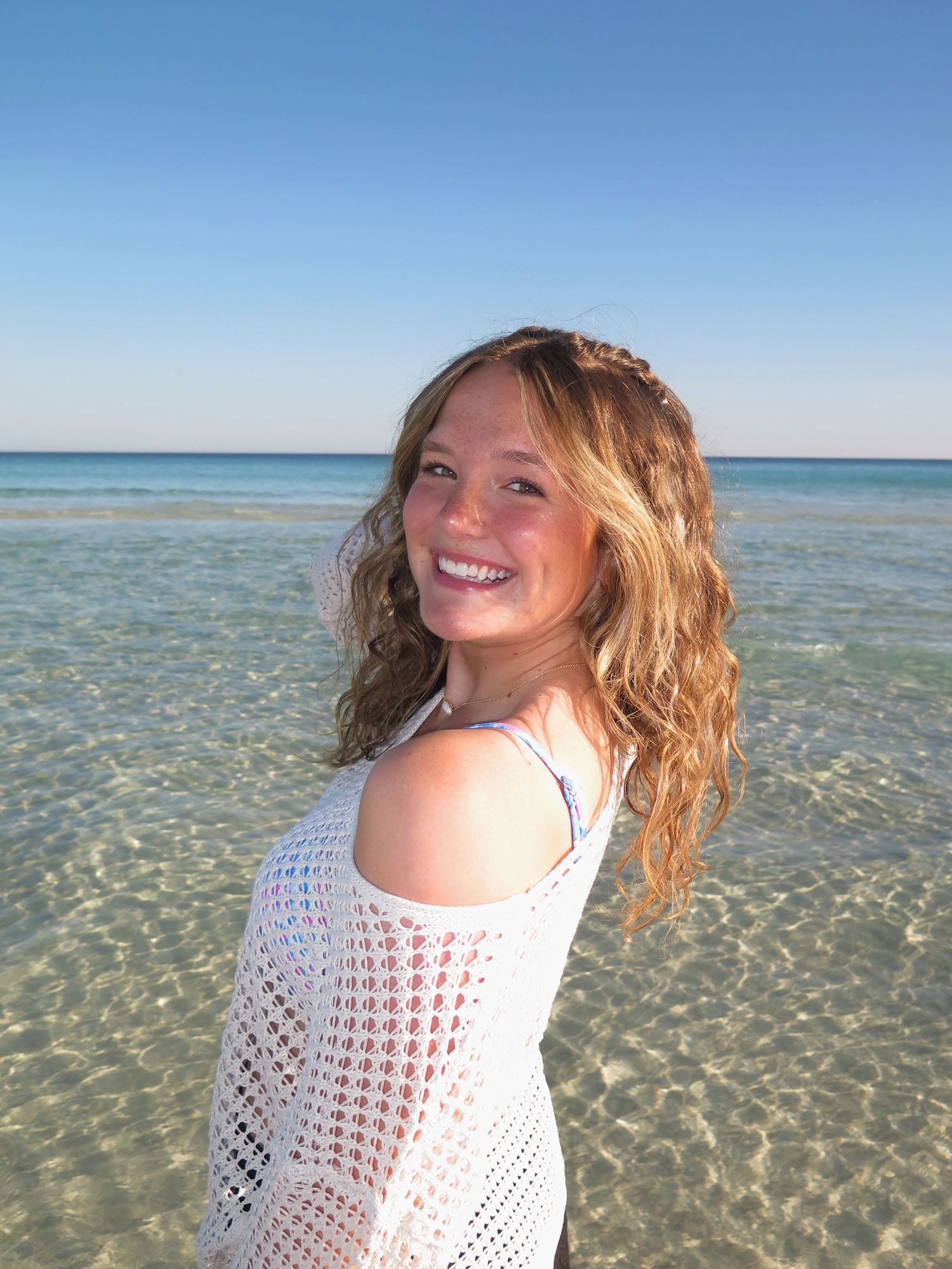 A smiling woman with wavy blonde hair looking back over her shoulder while standing in shallow, clear ocean water.