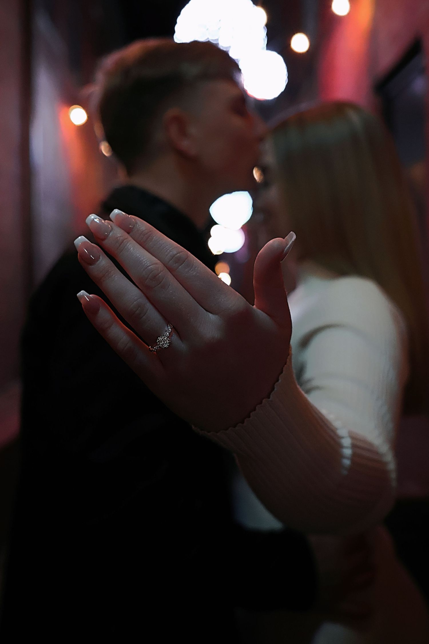 A man kissing a woman on the forehead while she places her hand on his chest, displaying an engagement ring.