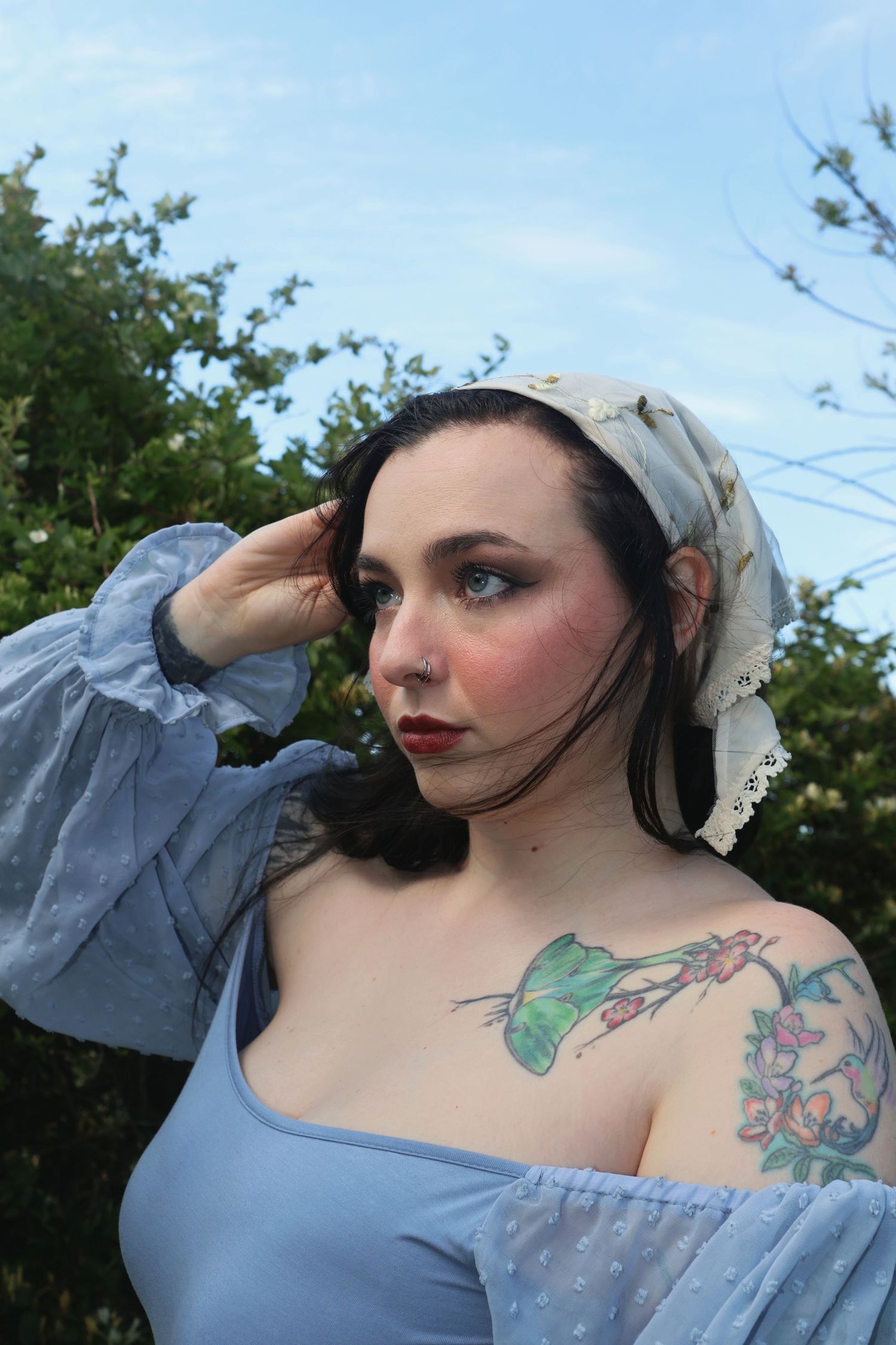 A woman wearing an embroidered bandana and off-the-shoulder blue top holding her hair as wind sweeps by.