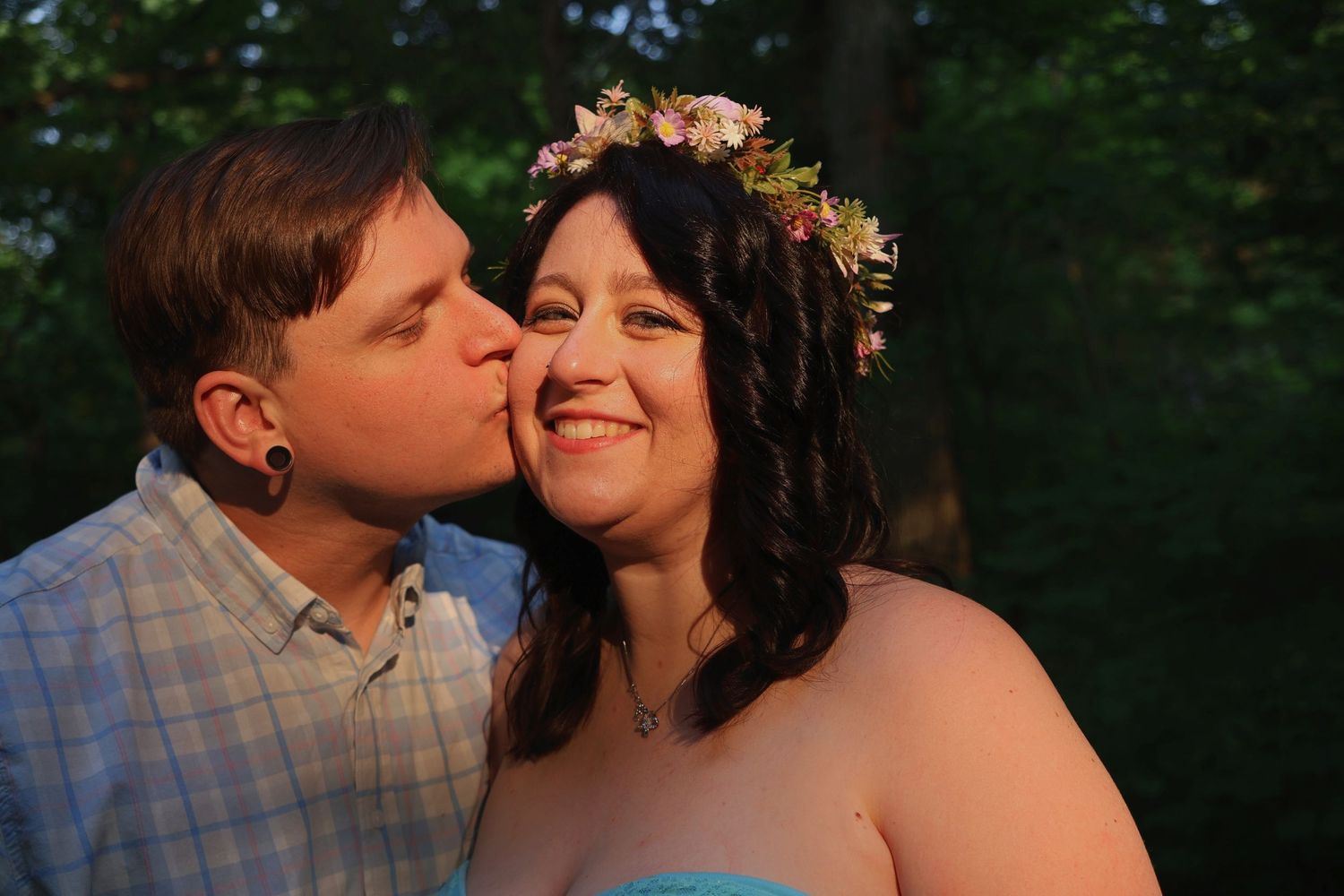 A man kissing a smiling woman wearing a pastel flower crown on the cheek outdoors during golden hour.