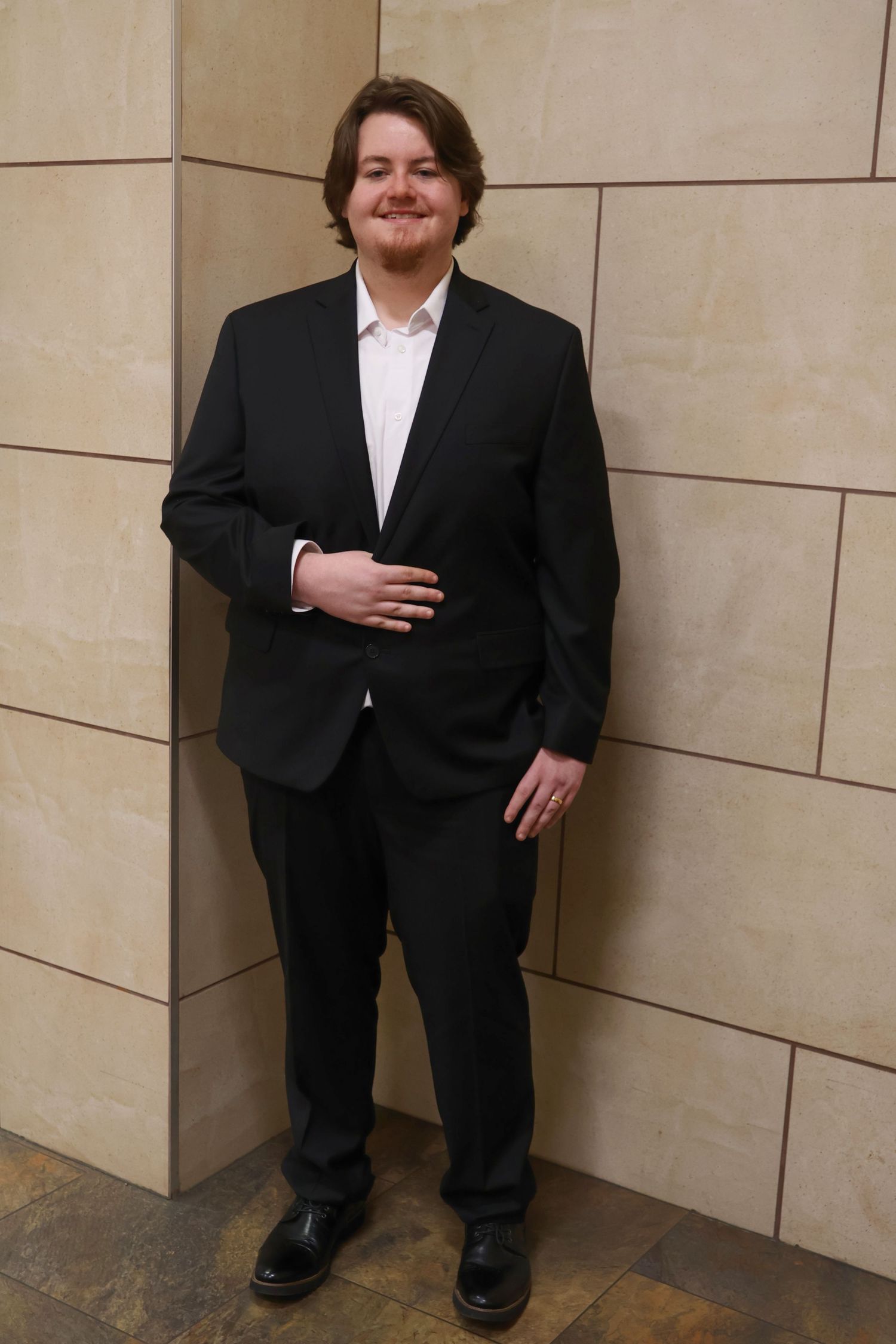 Full-length portrait of a smiling groom in a black suit resting his hand on his stomach against a tiled interior wall.