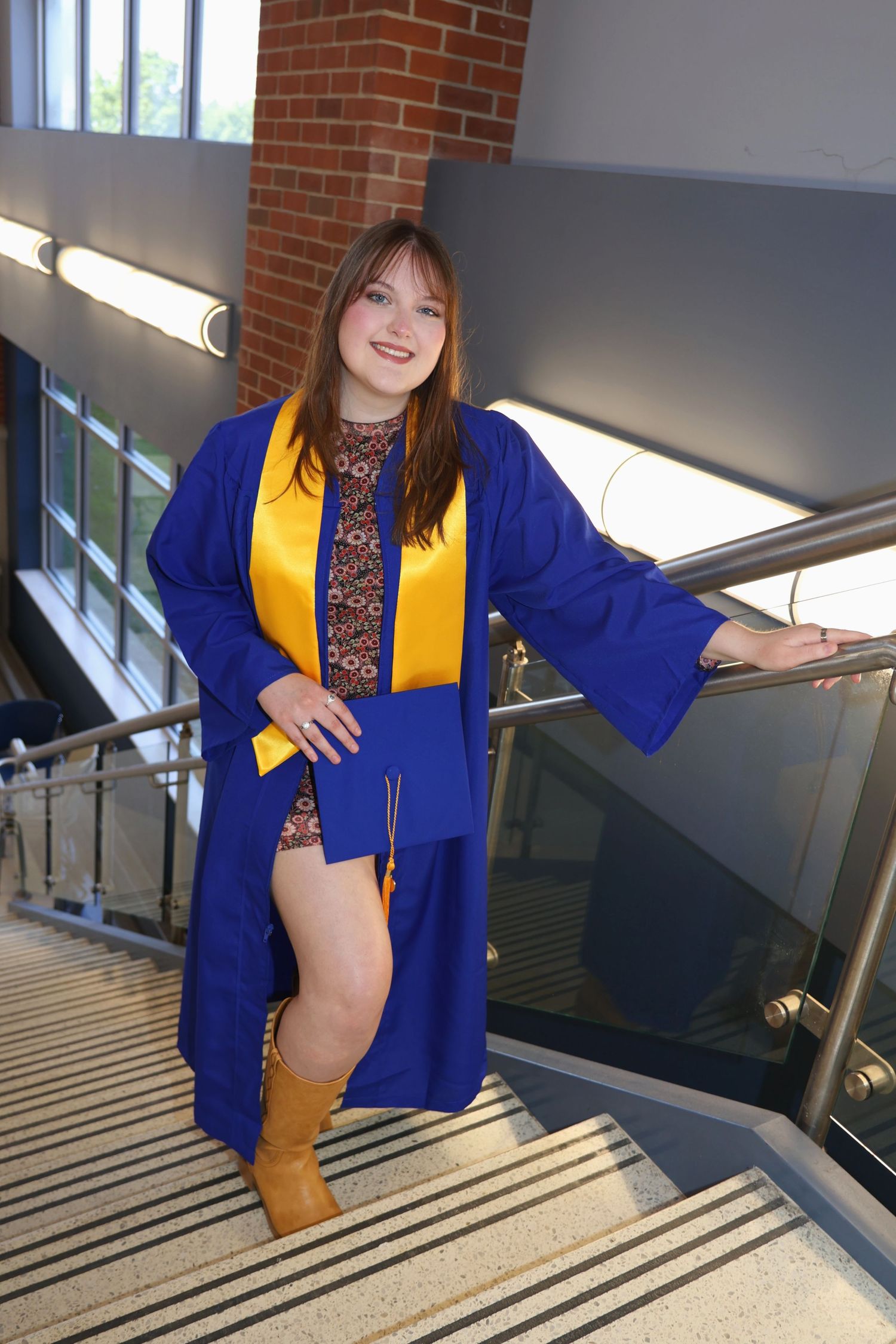 Full-length portrait of a woman in a blue graduation gown, yellow stole, and tan boots posing on an indoor staircase.