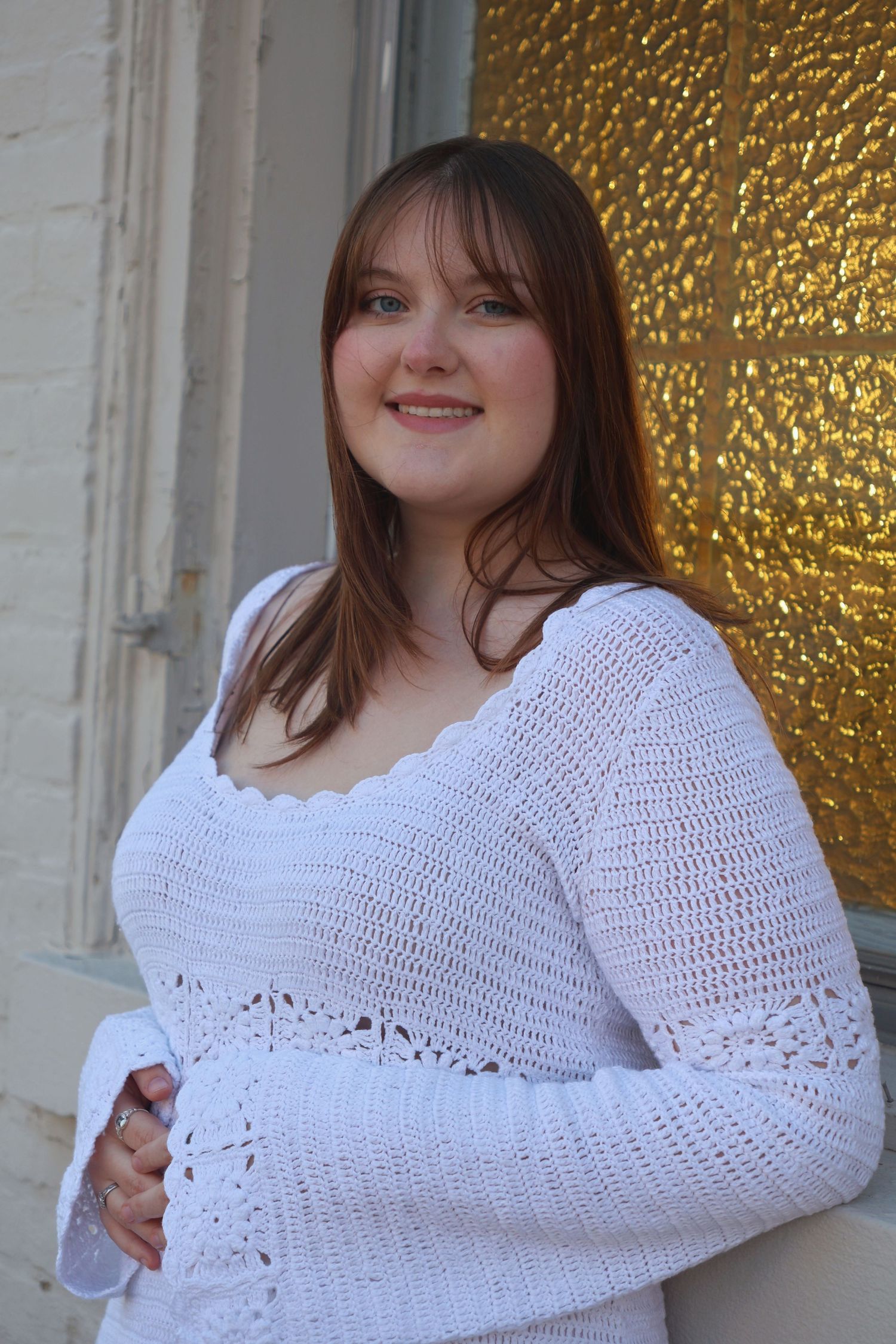 A smiling woman in a white knit long-sleeve dress leaning against a building entryway with a decorative glass door in the background.