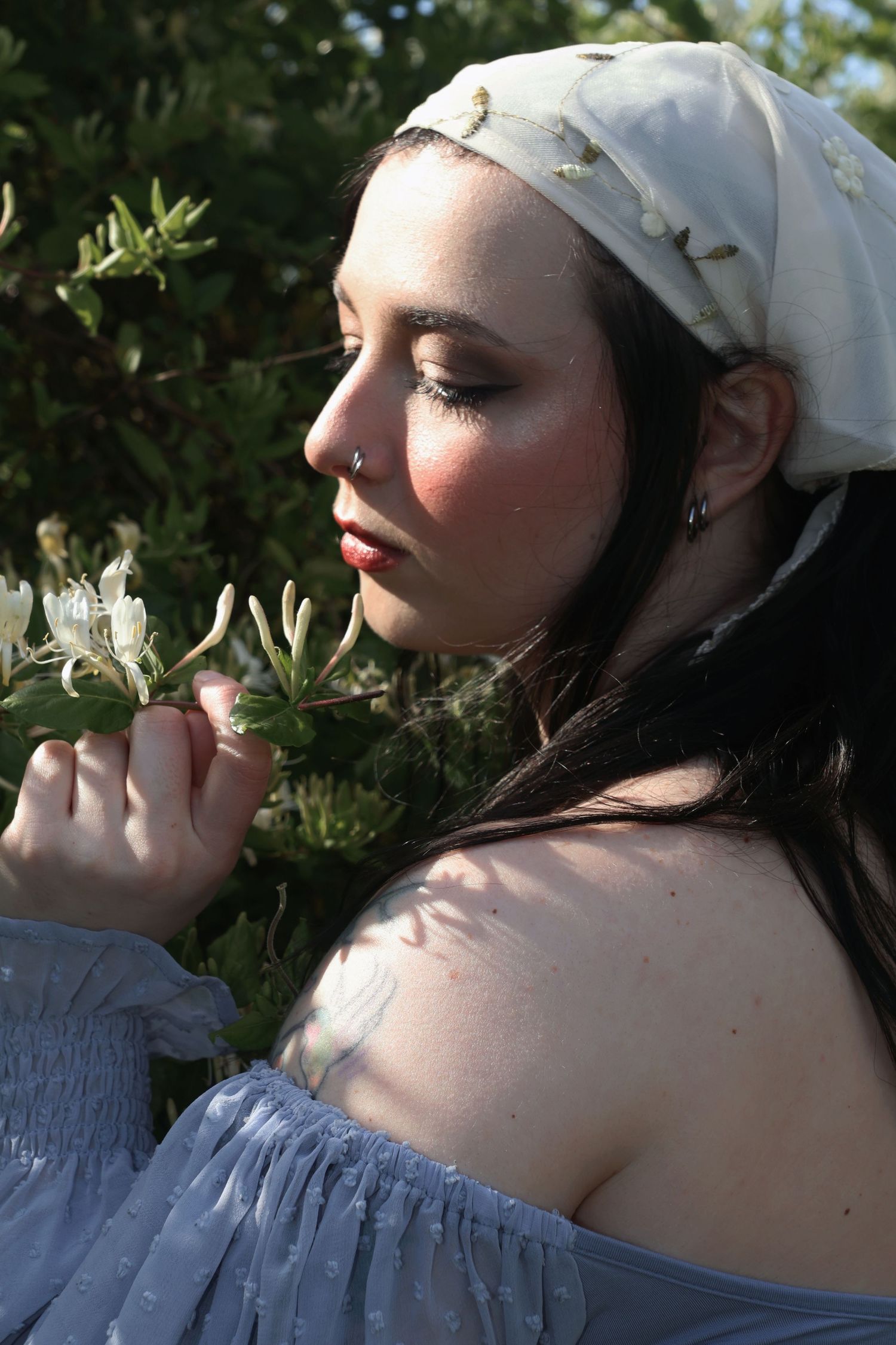 A woman wearing an embroidered bandana and off-the-shoulder blue top looking down at a honeysuckle branch.