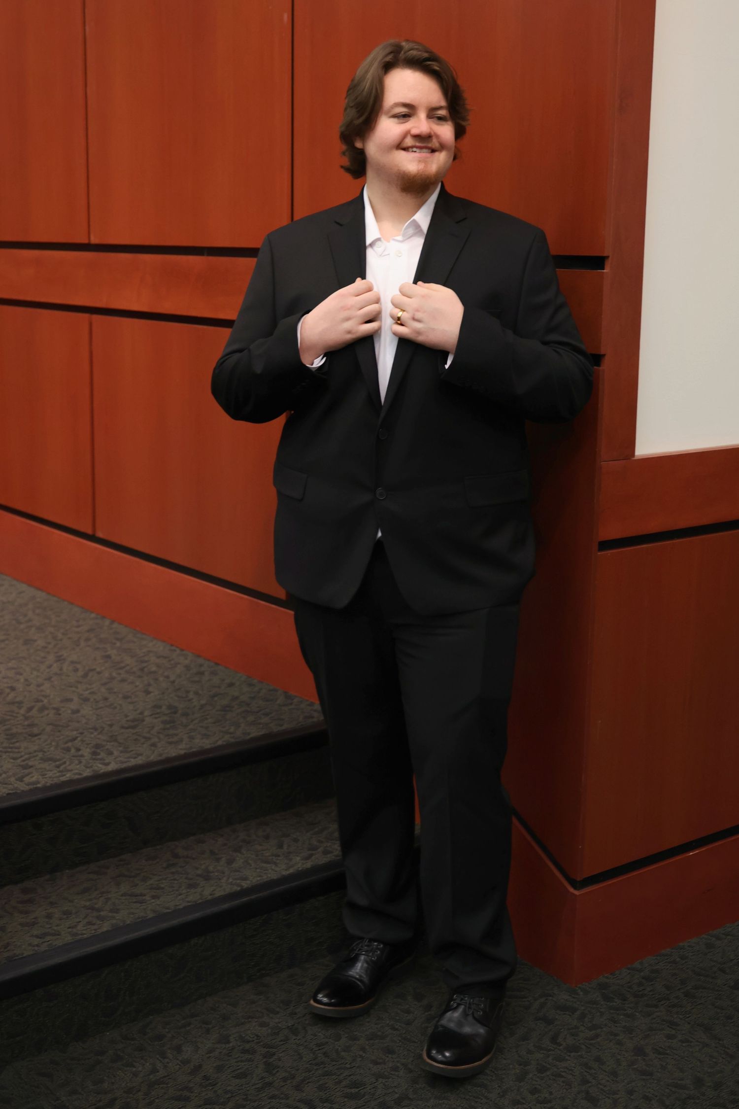 Full-length portrait of a smiling groom in a black suit adjusting his jacket lapels indoors near steps.