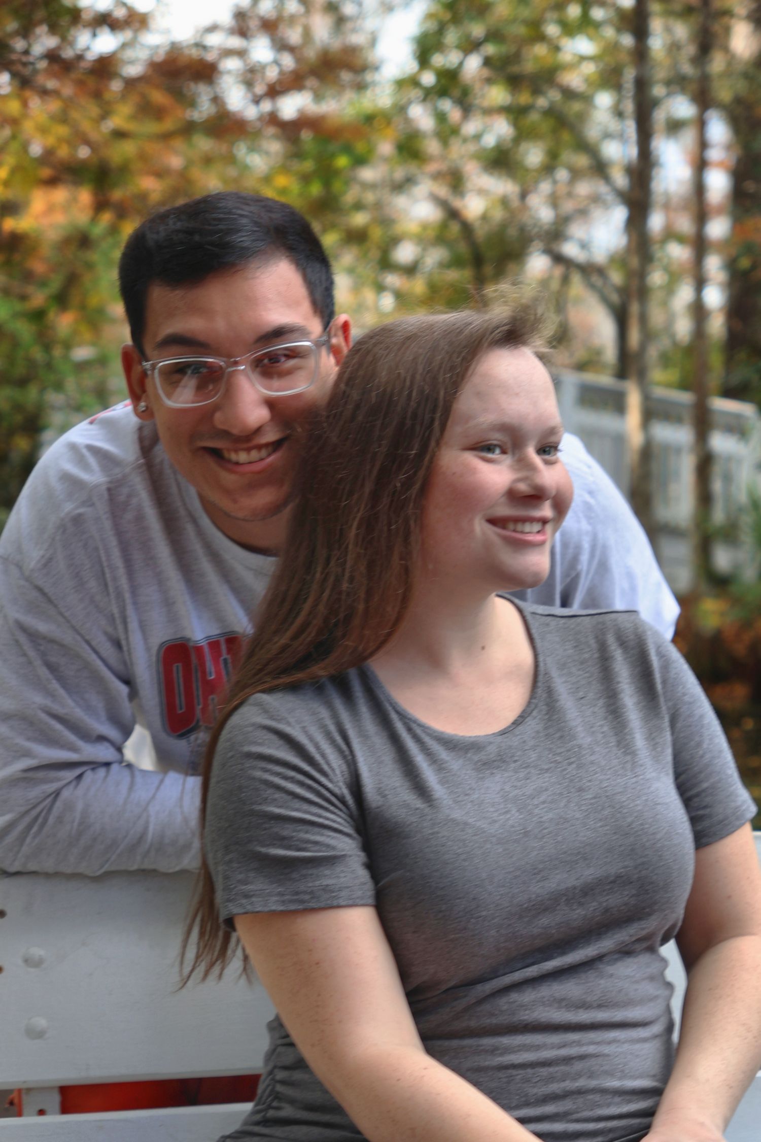 A smiling man leaning on a white fence behind a woman wearing a grey t-shirt, both looking off to the side outdoors.