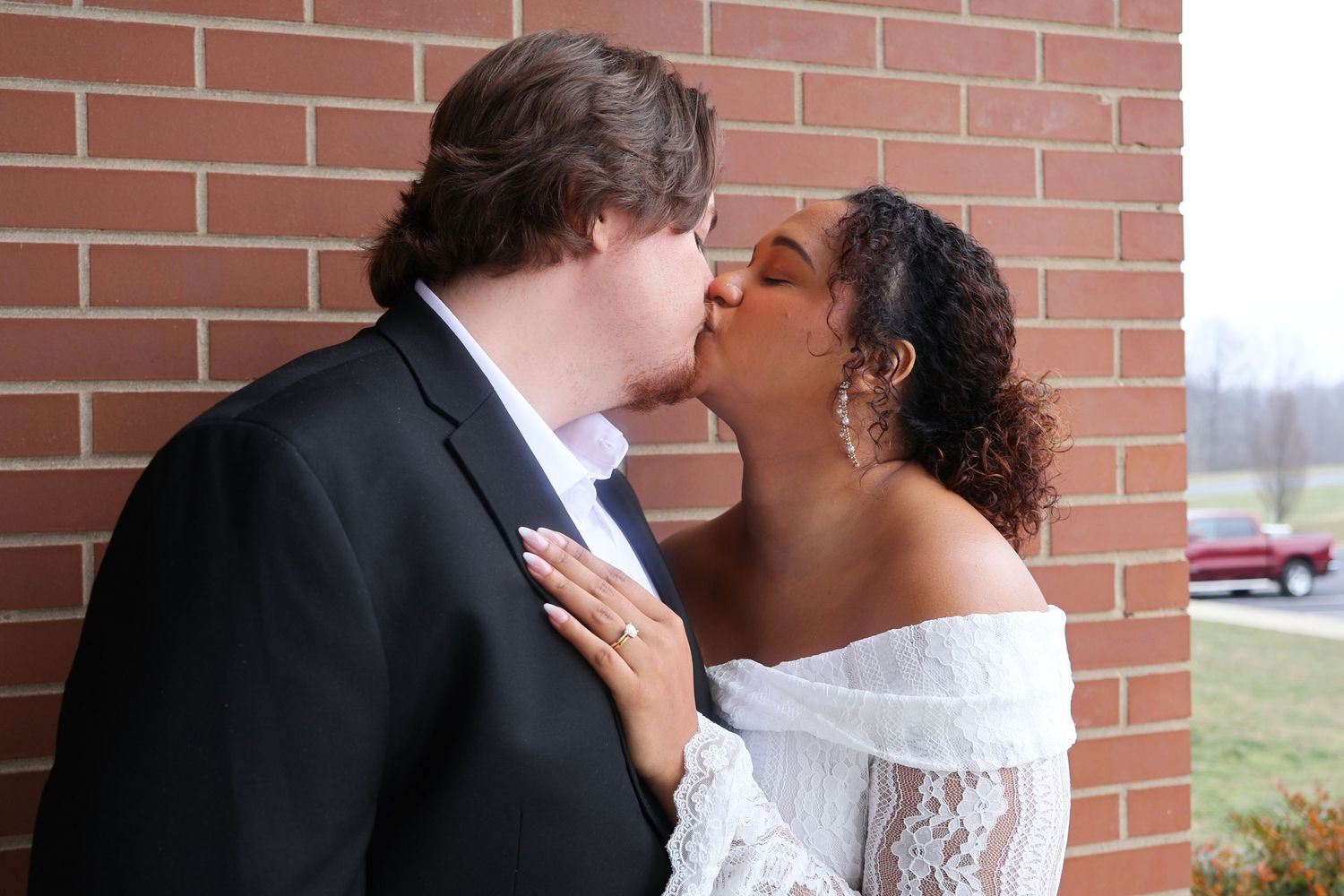 A newlywed couple kissing outside against a red brick wall, with the bride resting her hand on the groom's suit jacket.