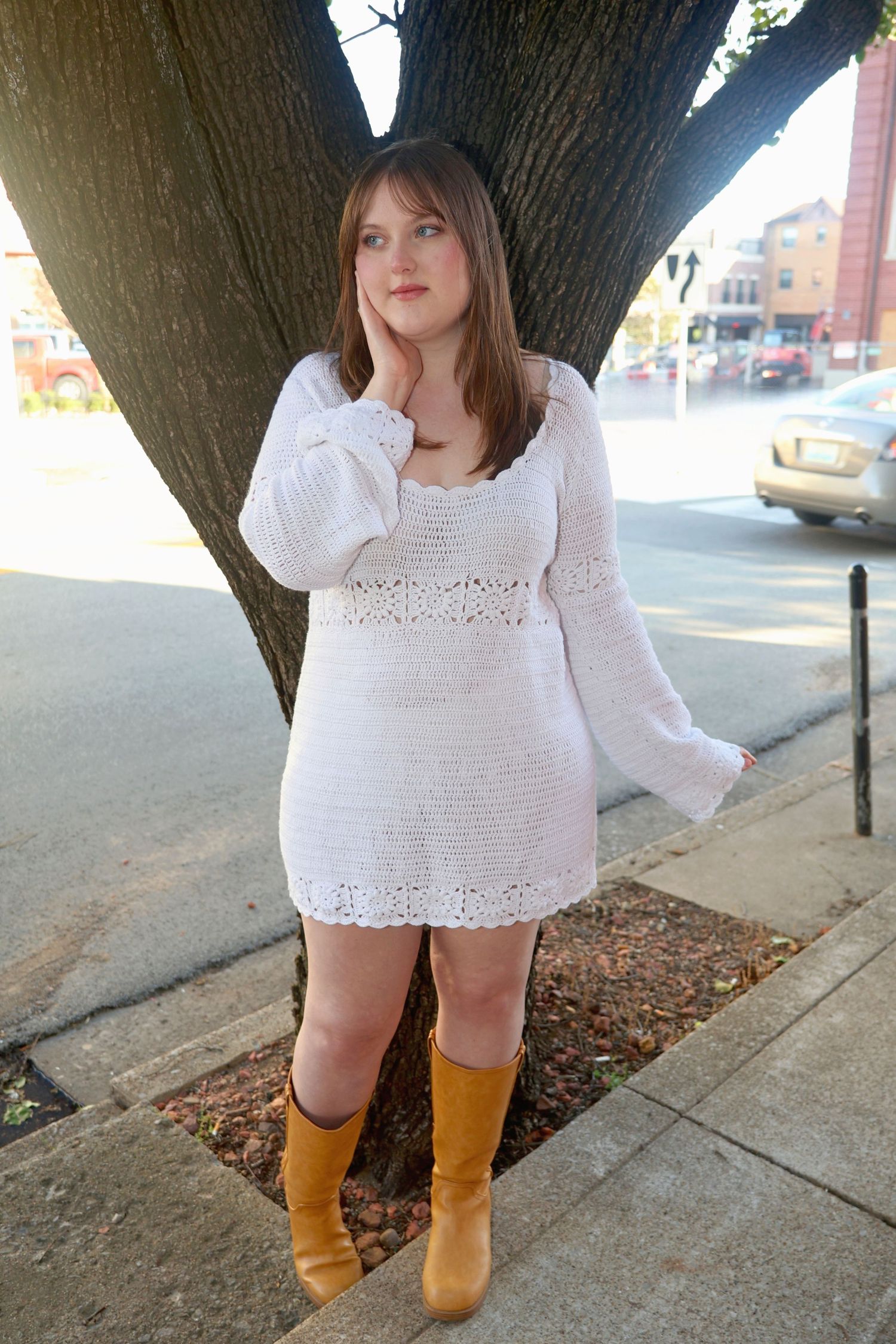 Full-length portrait of a woman in a white knit long-sleeve dress and tan boots leaning against a tree trunk along a city sidewalk.