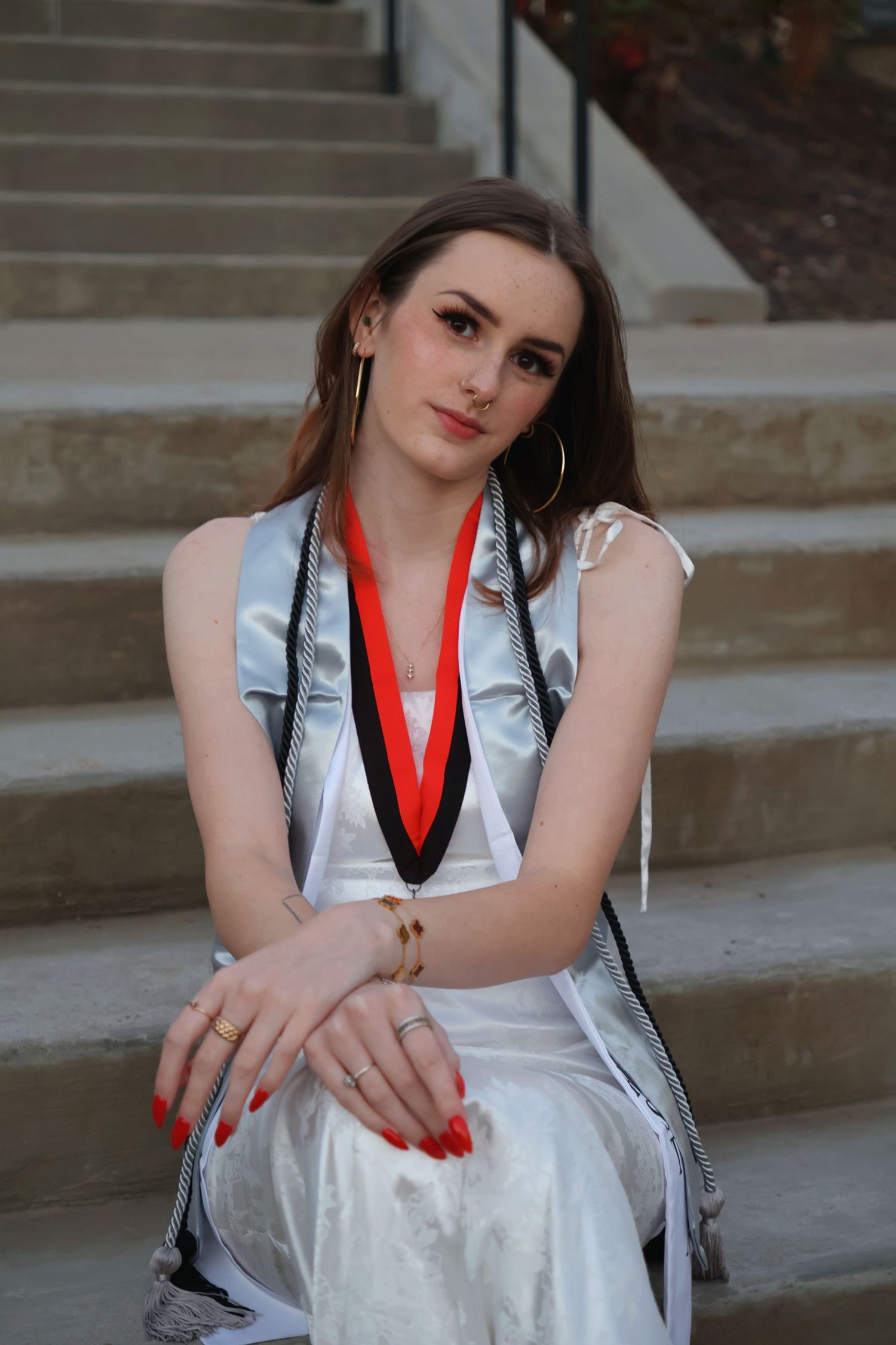 A graduating woman sitting on outdoor concrete steps in a white dress, stole, and honor cords, leaning forward with her hands on her knees