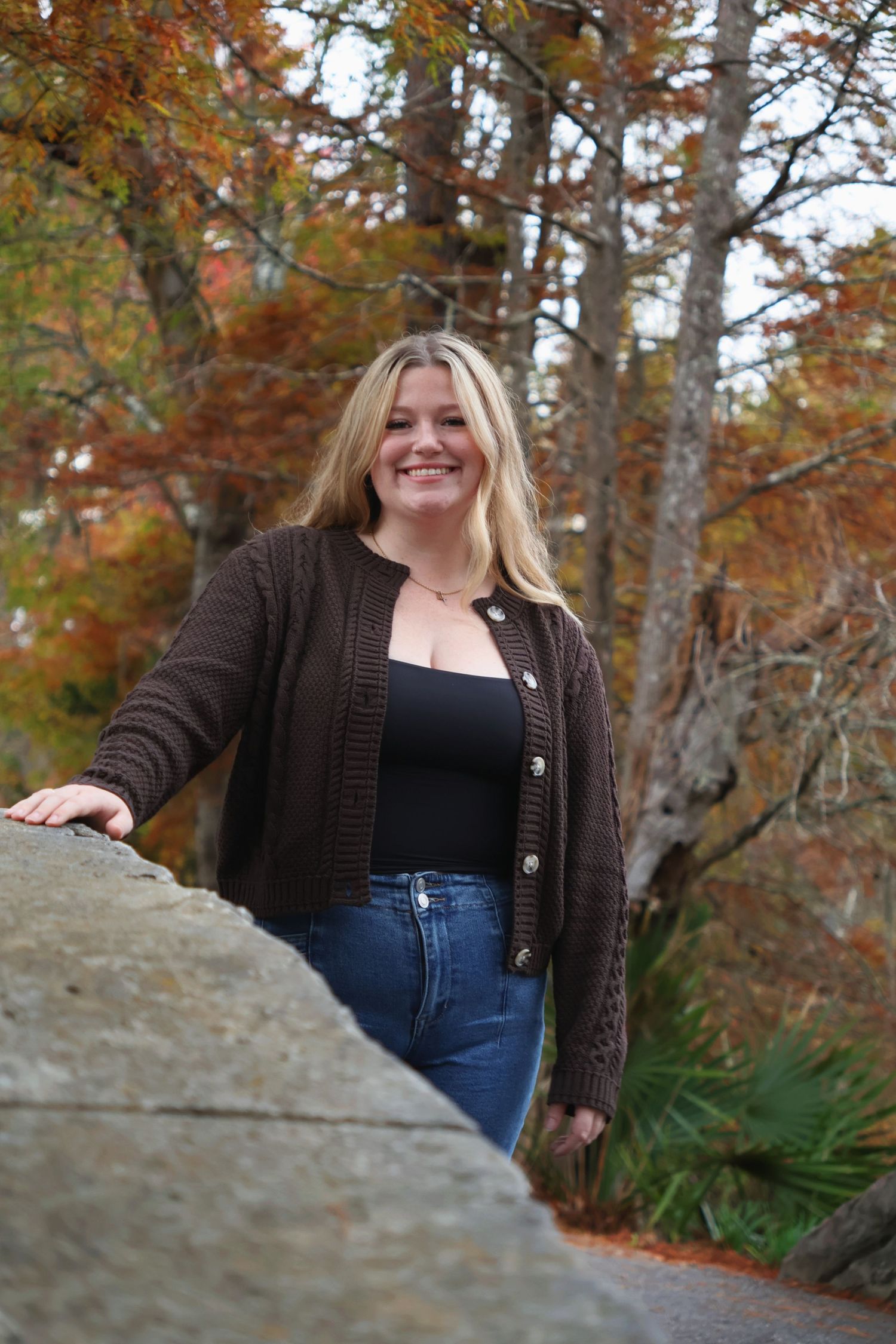 A smiling woman in a brown knit cardigan standing on a stone bridge with fall foliage behind her.