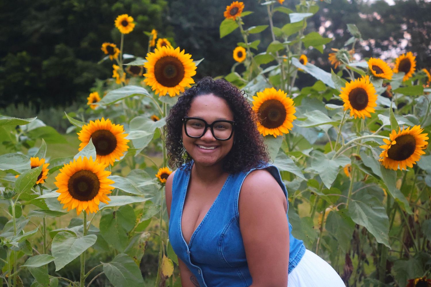 A woman  wearing glasses, a denim top standing in a sunflower field smiling wide.