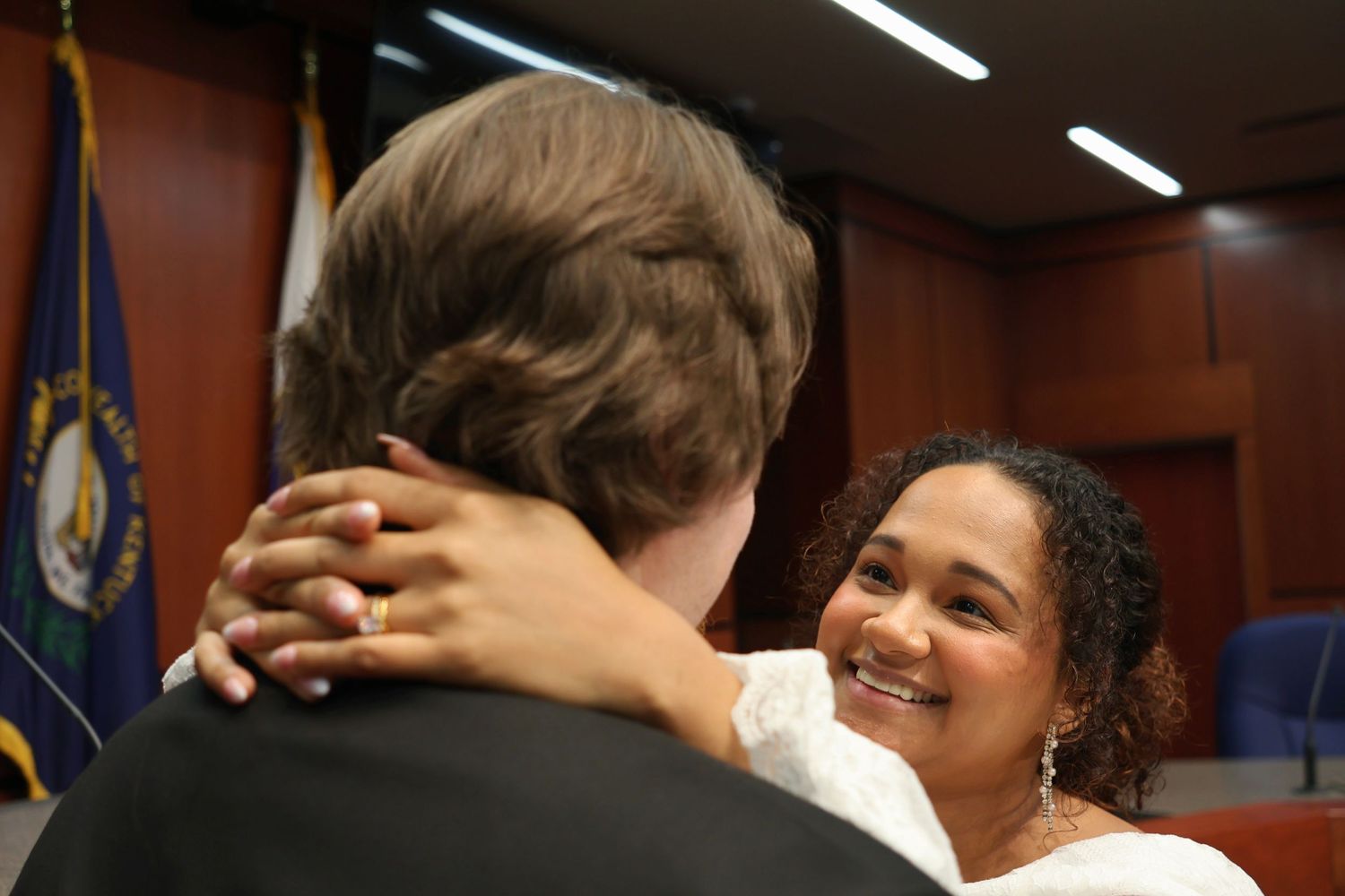 A smiling bride hugging her groom around the neck inside a courtroom setting with flags from a court house sit in the background.