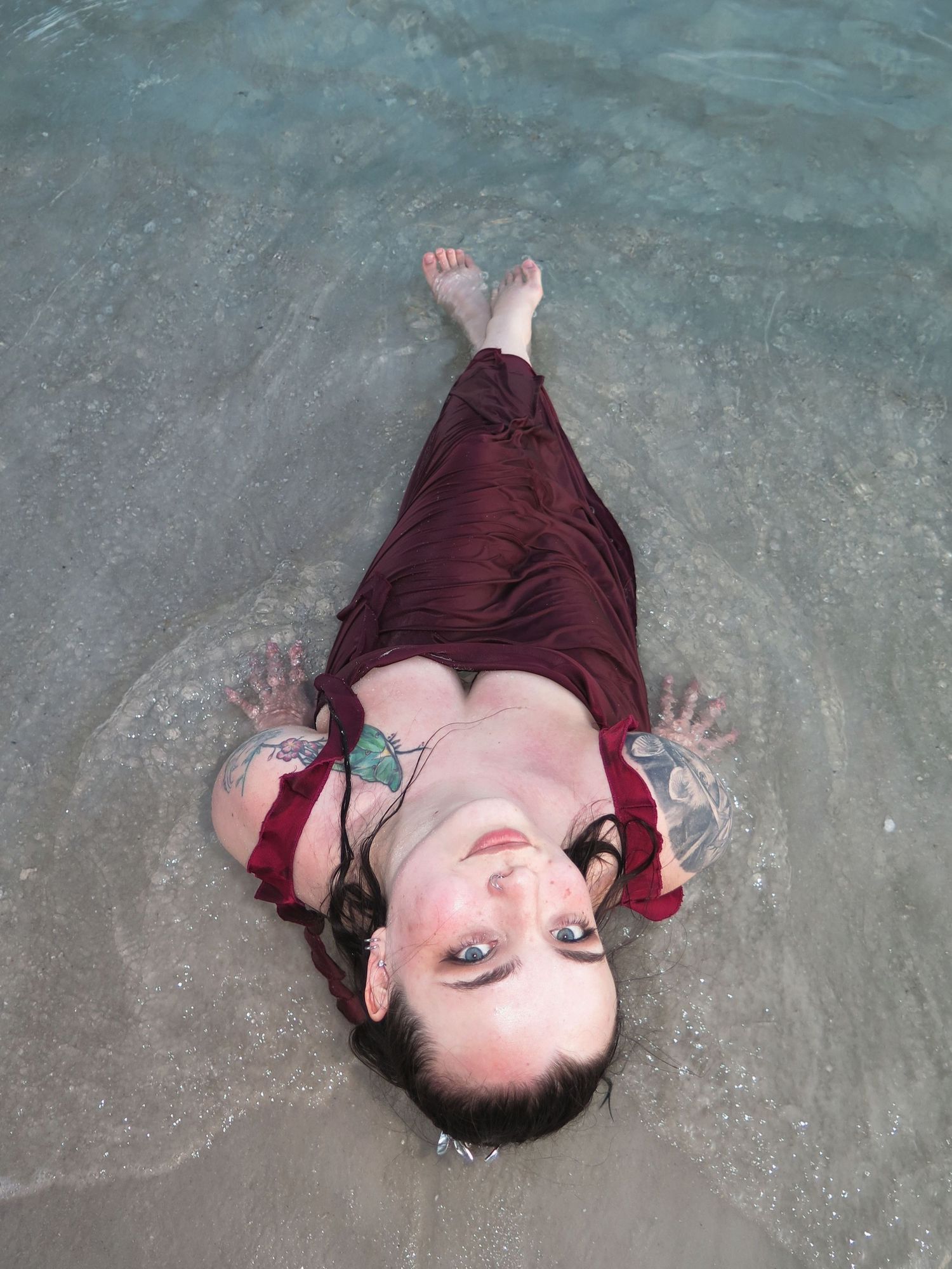 A woman with wet hair wearing a soaked maroon dress lying in shallow ocean water, looking up at the camera with a soft smile.