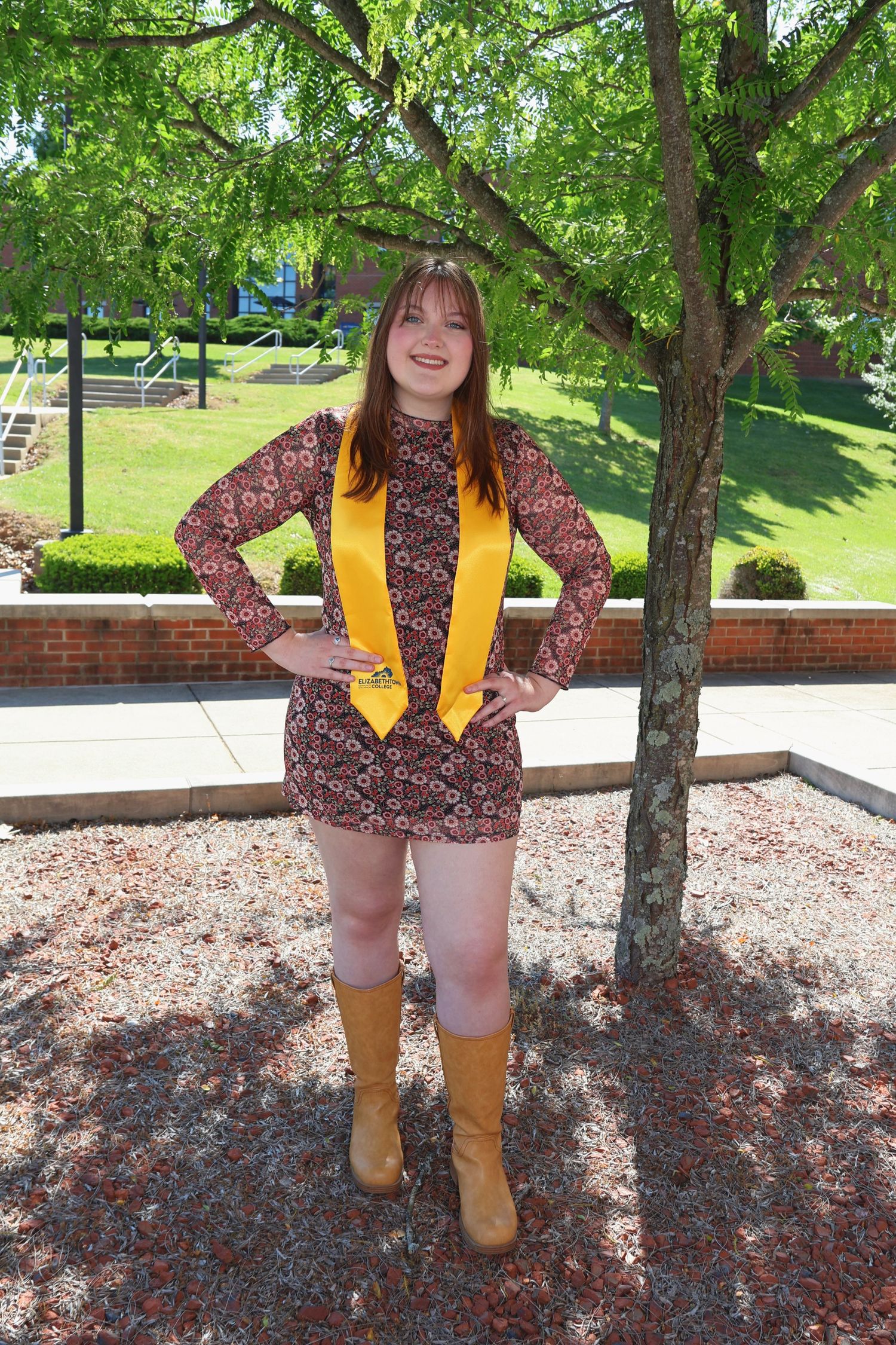 Full-length portrait of a woman standing outdoors near a tree with her hands on her hips, wearing a yellow graduation stole and tall boots