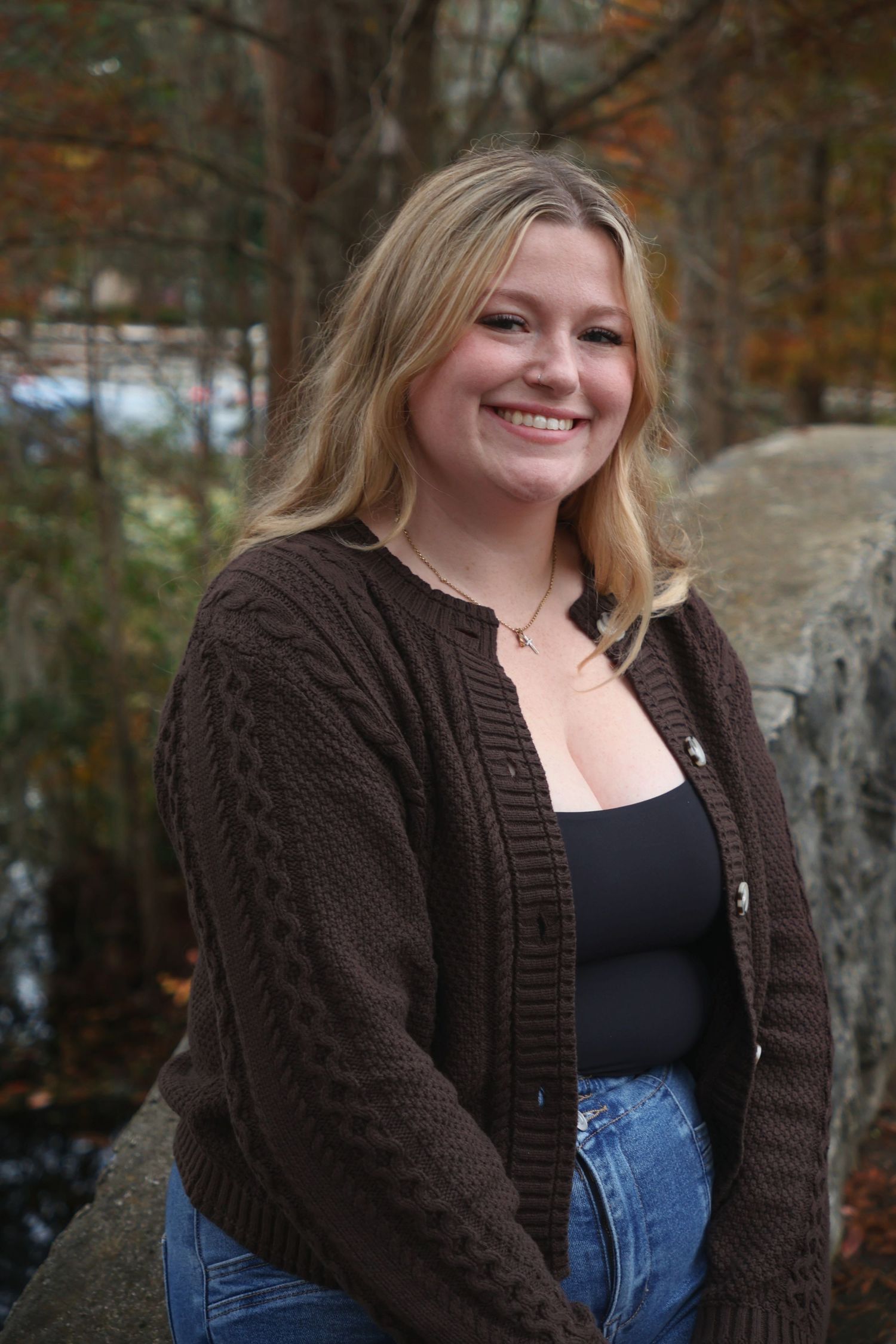 A smiling woman wearing a brown knit cardigan over a dark top, posing near a stone structure with fall foliage behind her.