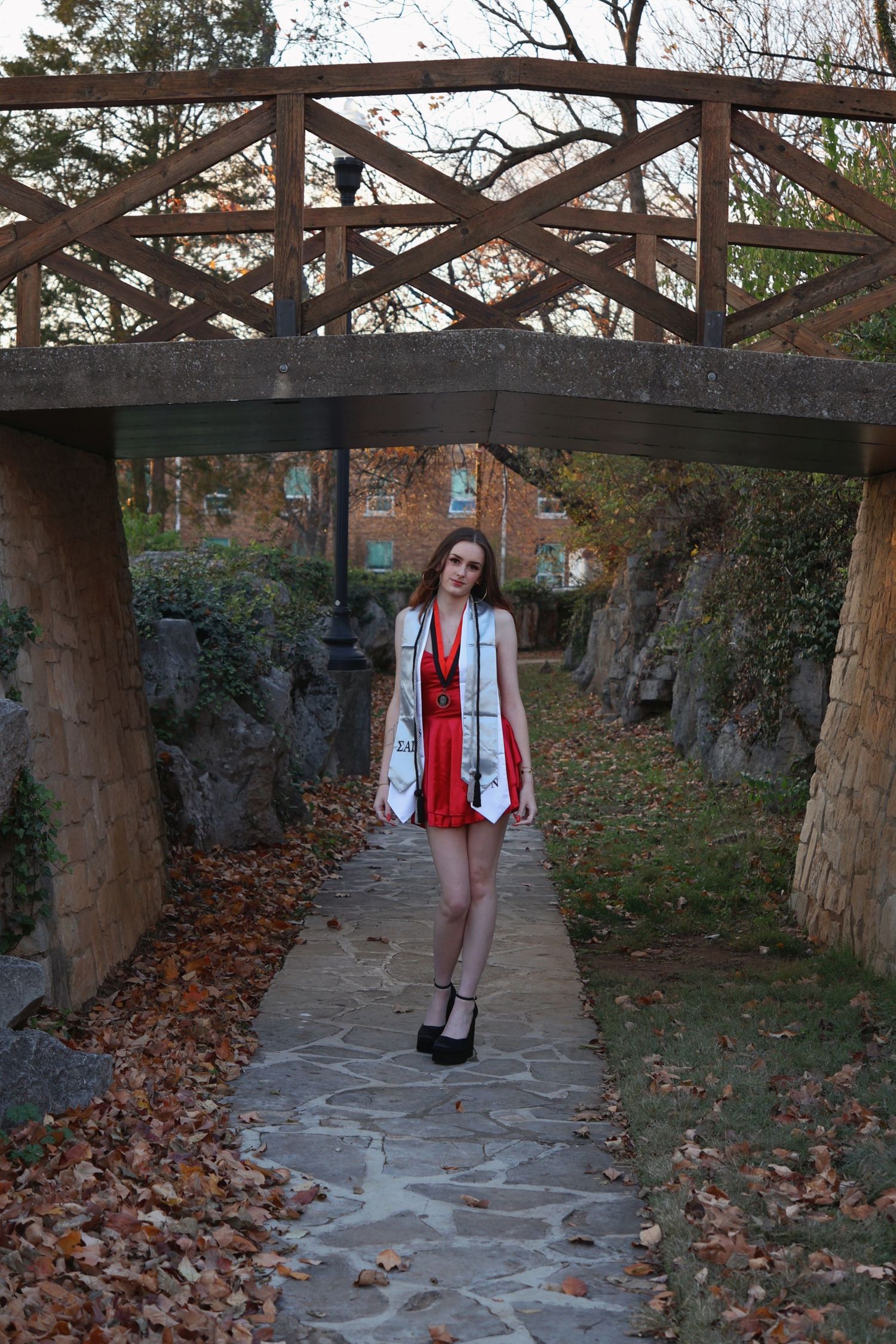Full-body portrait of a woman graduate in a red dress and stole standing on a stone pathway beneath a wooden footbridge.
