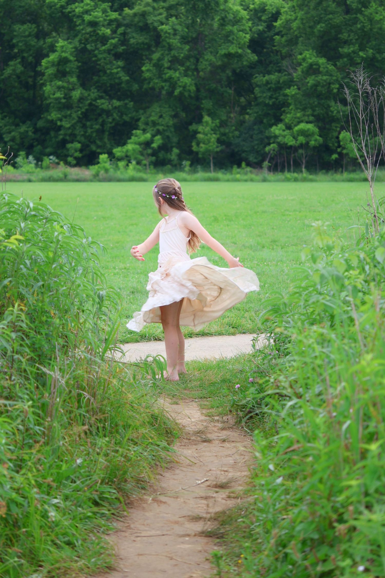 A girl in a light-colored dress twirling along a dirt path through a grassy field with trees in the distance.