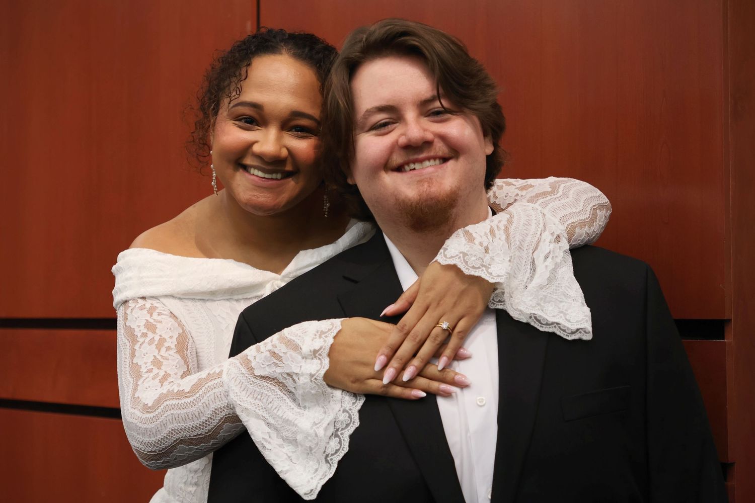 A smiling bride with her arm wrapped around the shoulder of her groom as they pose together against a wood-paneled wall.
