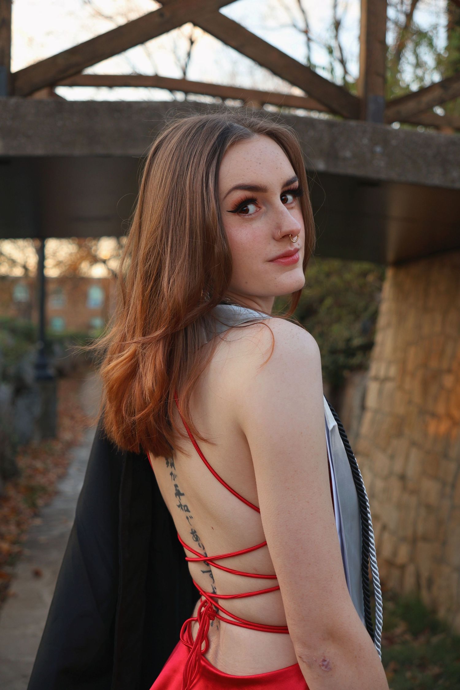 A woman looking back over her shoulder in a strappy backless red dress near a wooden bridge wearing a graduation stole and holding her gown.