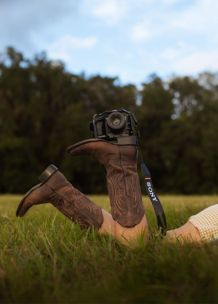 A picture of a female holding a camera with her boots in a green field