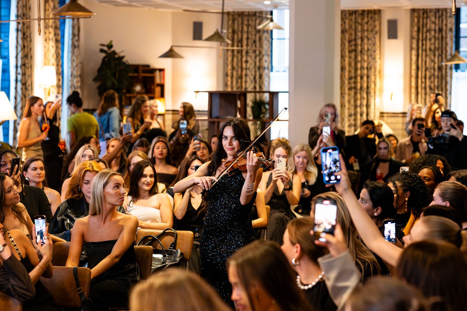Ten women event guests raise their arms in celebration on a crowded dance floor at an indoor venue.