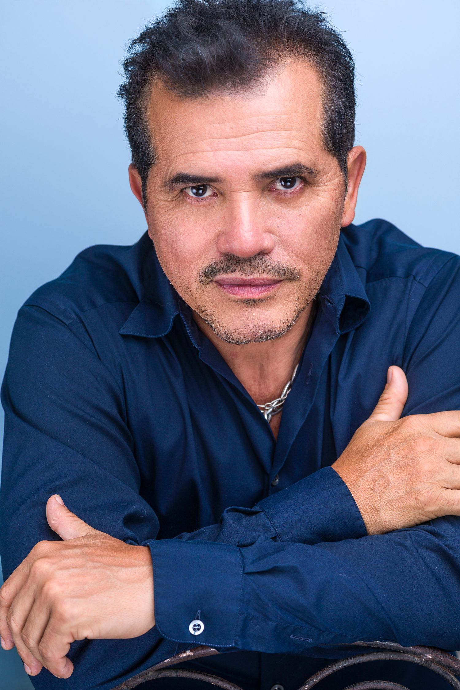 Middle-aged Latino man in a dark blue shirt with arms crossed leaning forward against a light blue background.