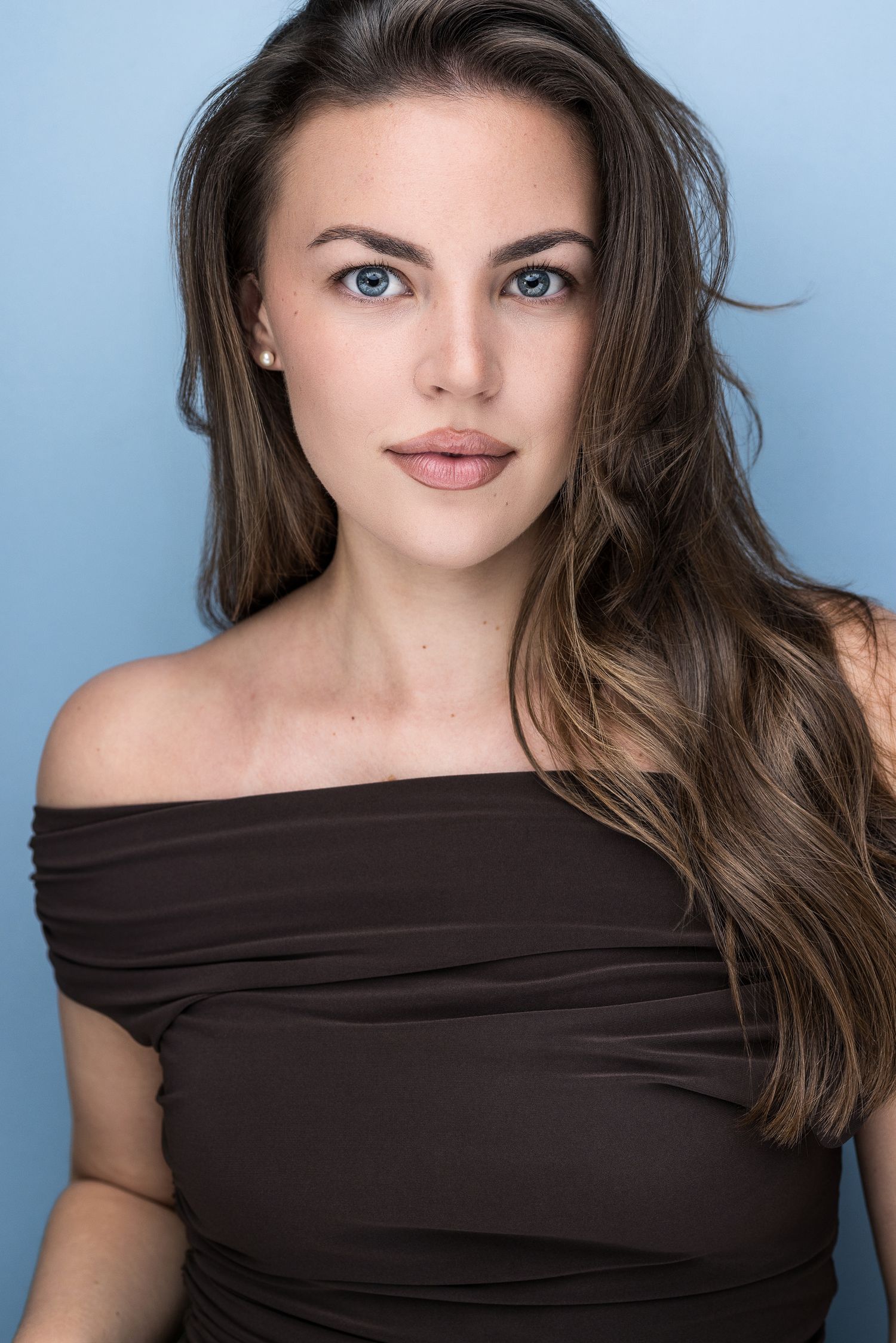 A young woman with long brunette hair and blue eyes wearing a brown off-shoulder top against a blue background.