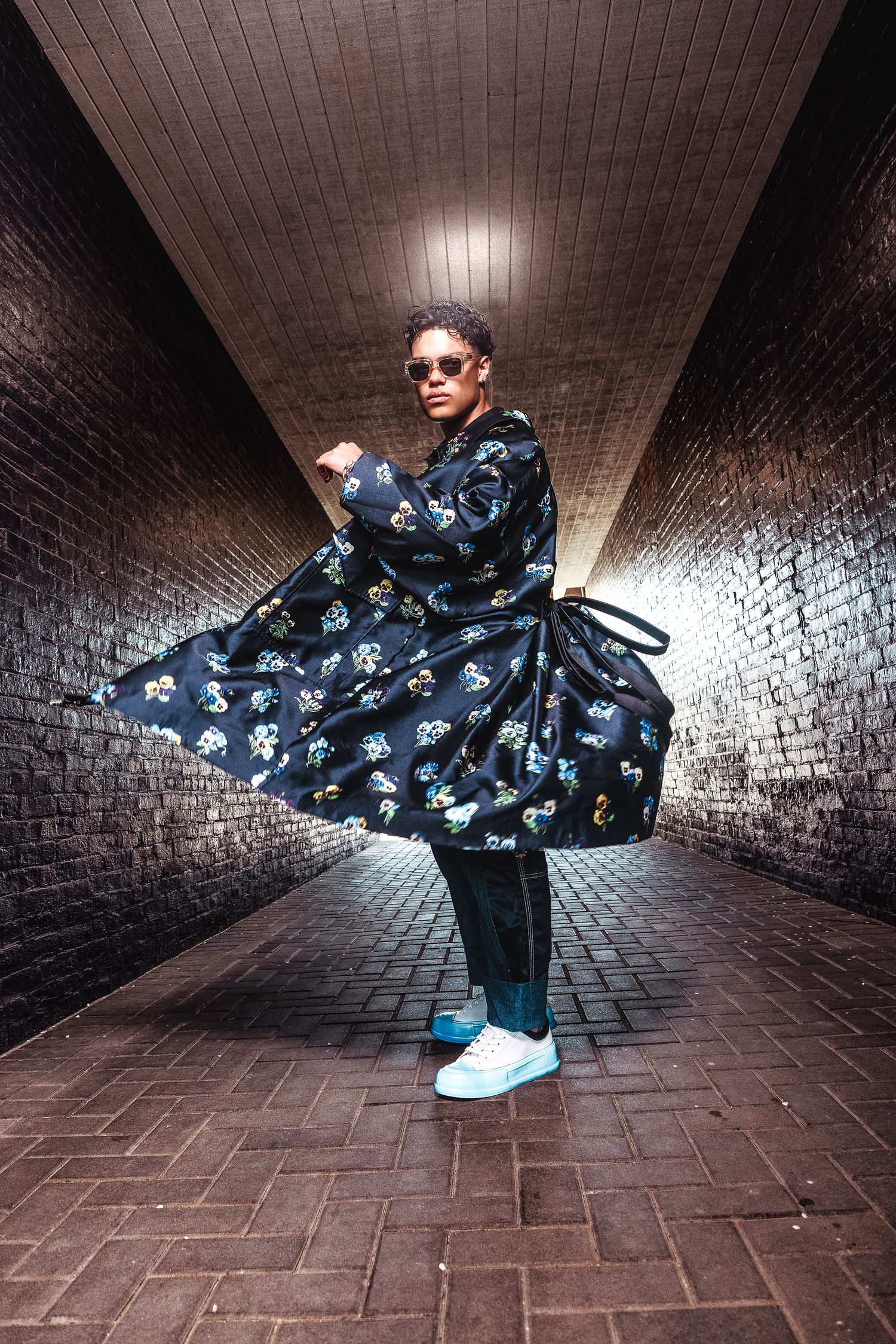 Person in floral printed coat posing in brick tunnel with blue sneakers.