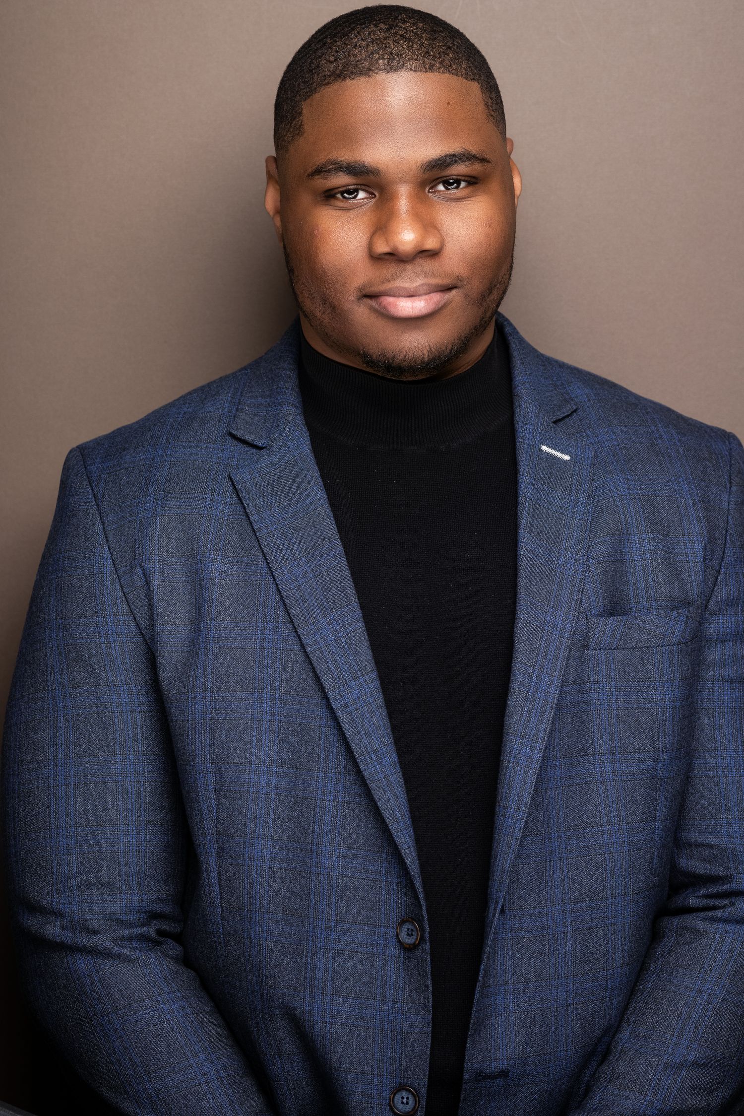 Professional headshot of a young man in a blue plaid blazer and black turtleneck against a gray background.