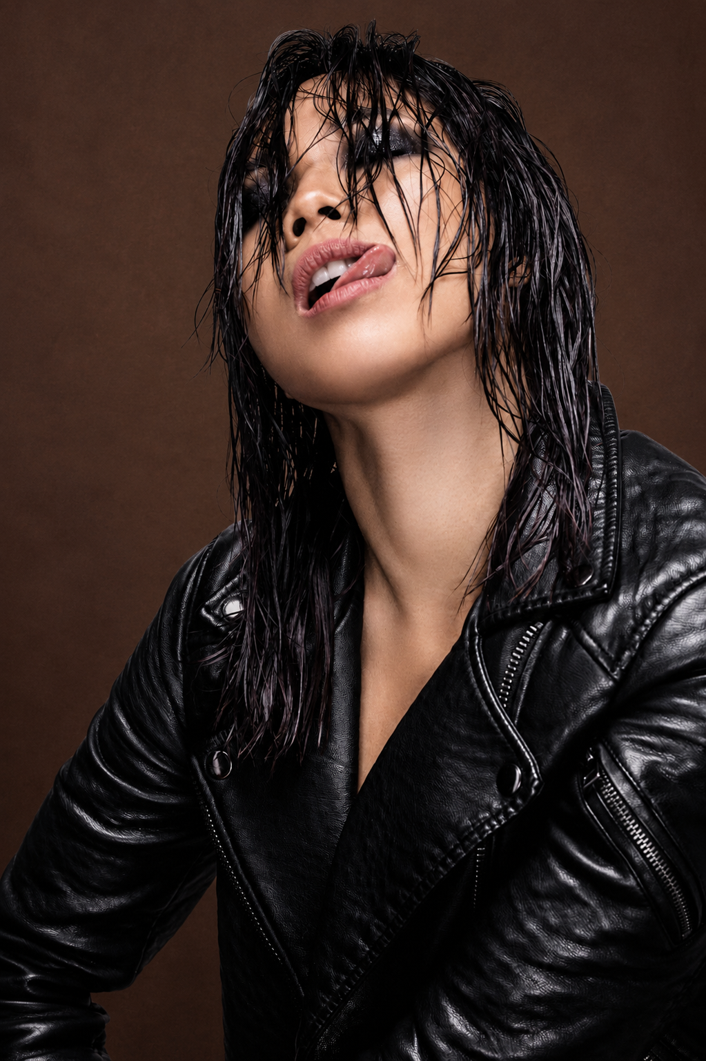 Woman in black leather jacket with wet hair posing dramatically against dark brown background.