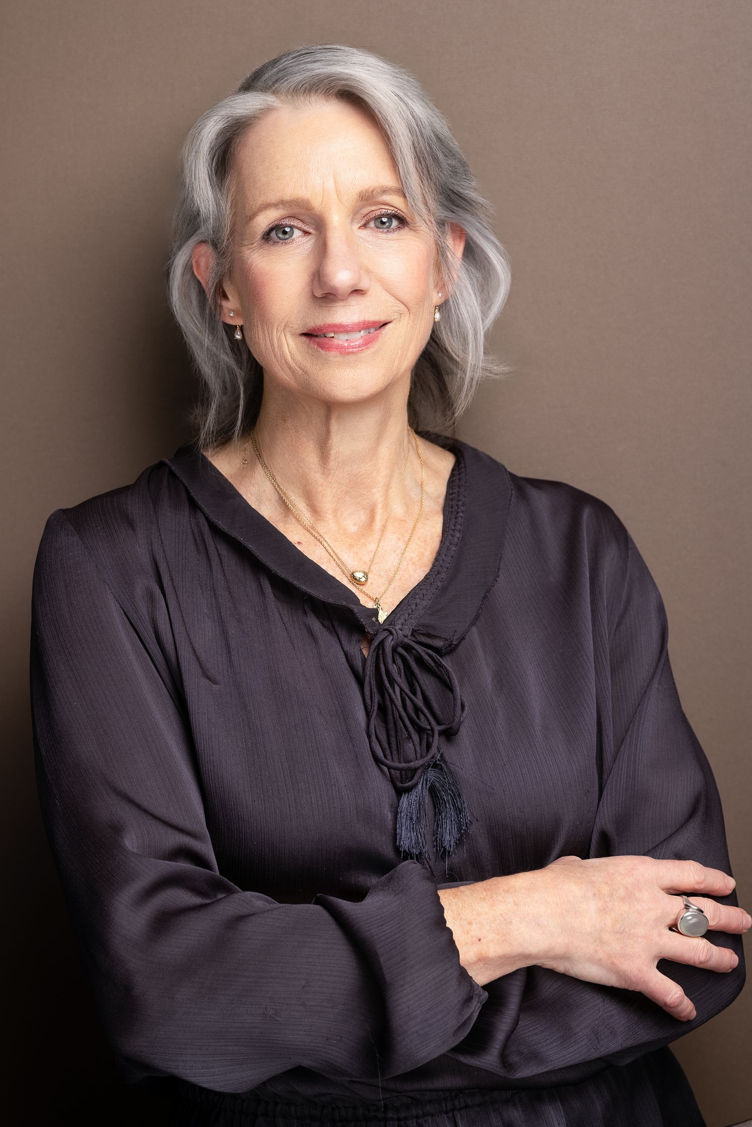 Elegant older woman with silver hair in dark blouse, arms crossed, wearing jewelry, professional headshot.
