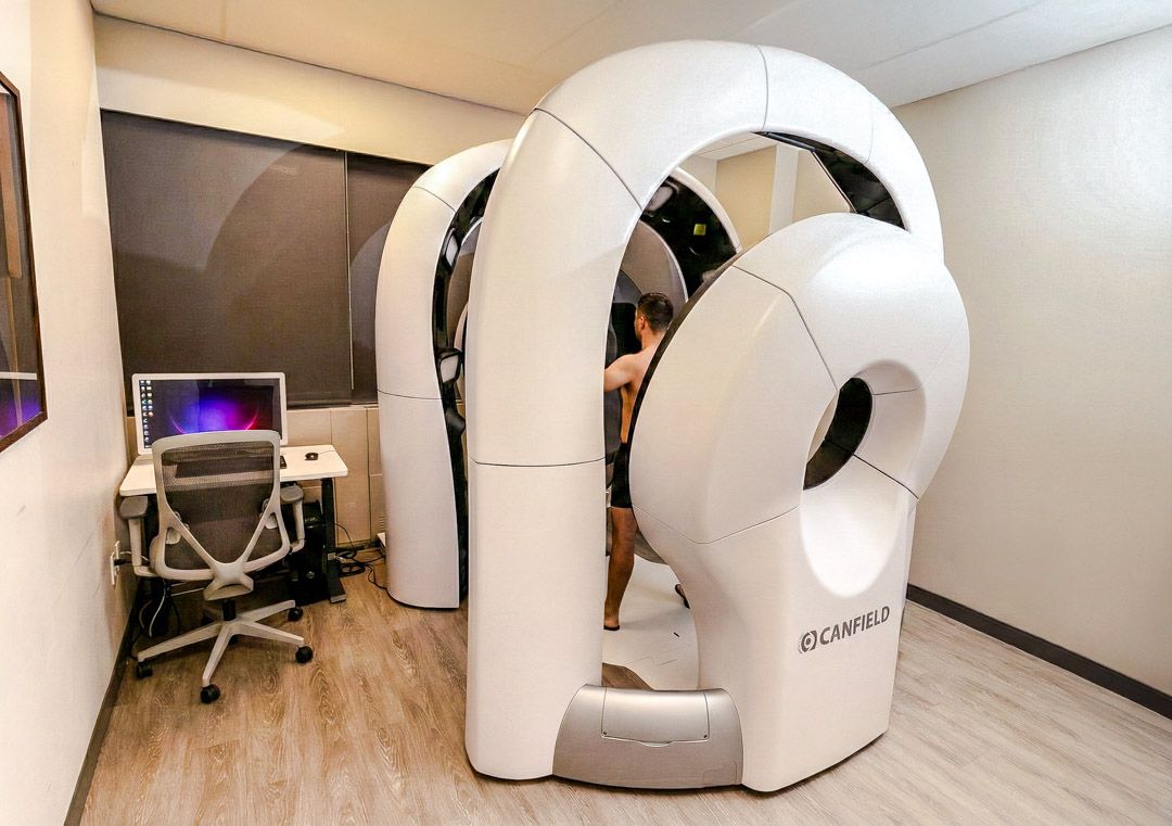 Person standing inside a white Fit3D-style full-body 3D scanner in a clean room with a desk, computer monitor and office chair nearby.