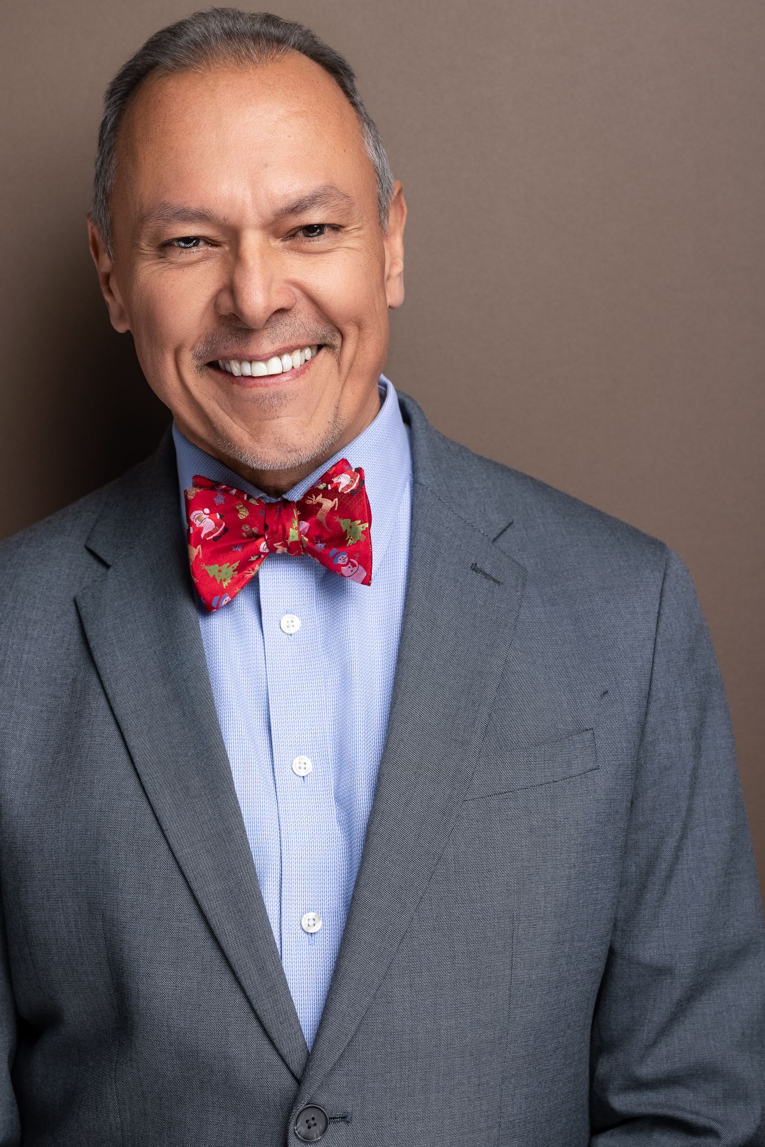 Professional headshot of a smiling man in a gray suit with a red floral bow tie against a brown background.