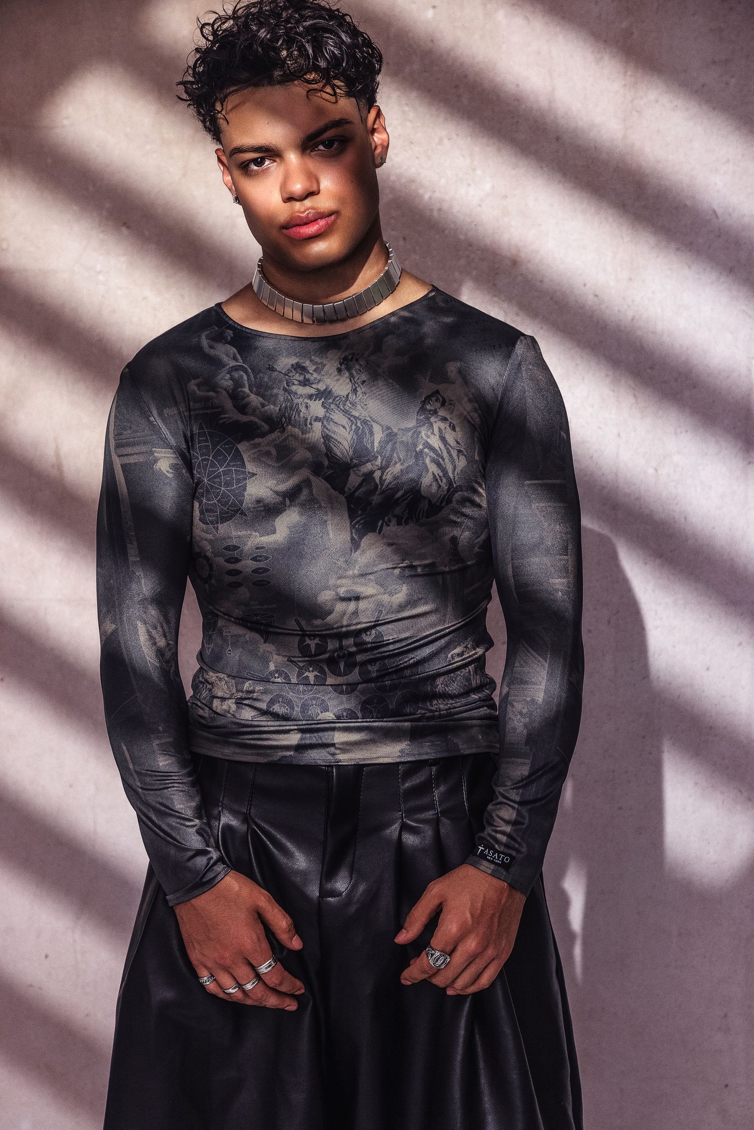 Young man in dark graphic long-sleeve top and black satin skirt, wearing a choker necklace, posing against a textured wall.