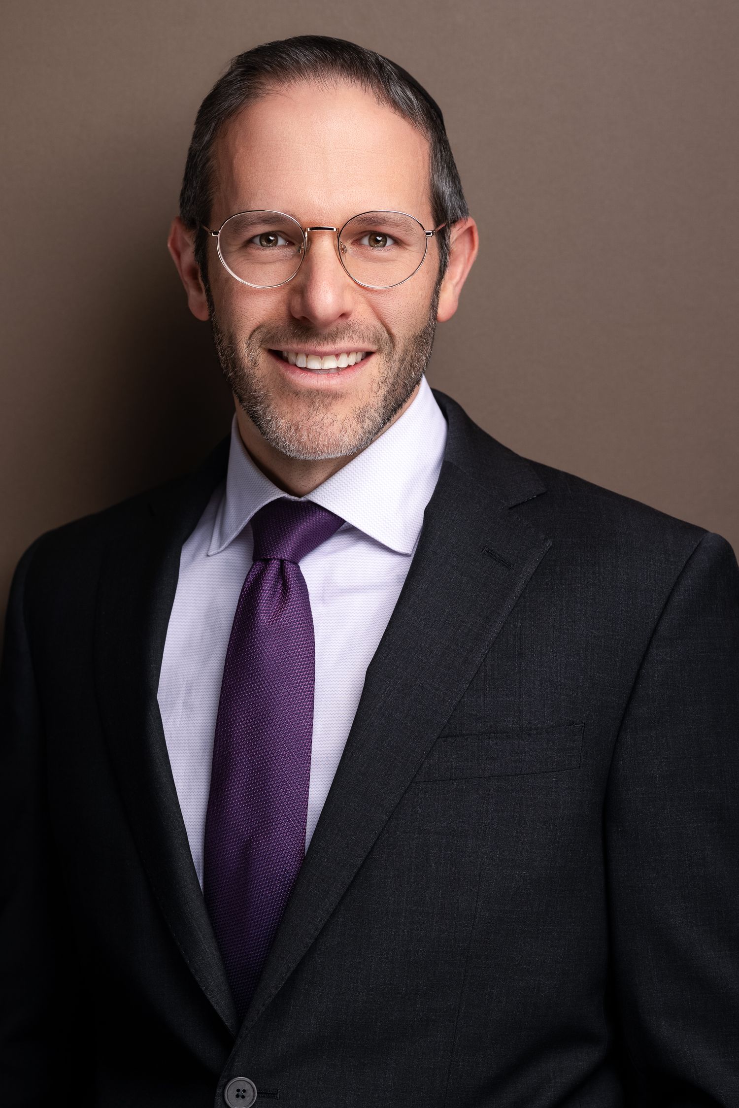 Professional man in dark suit and purple tie with round glasses smiling confidently against brown background.