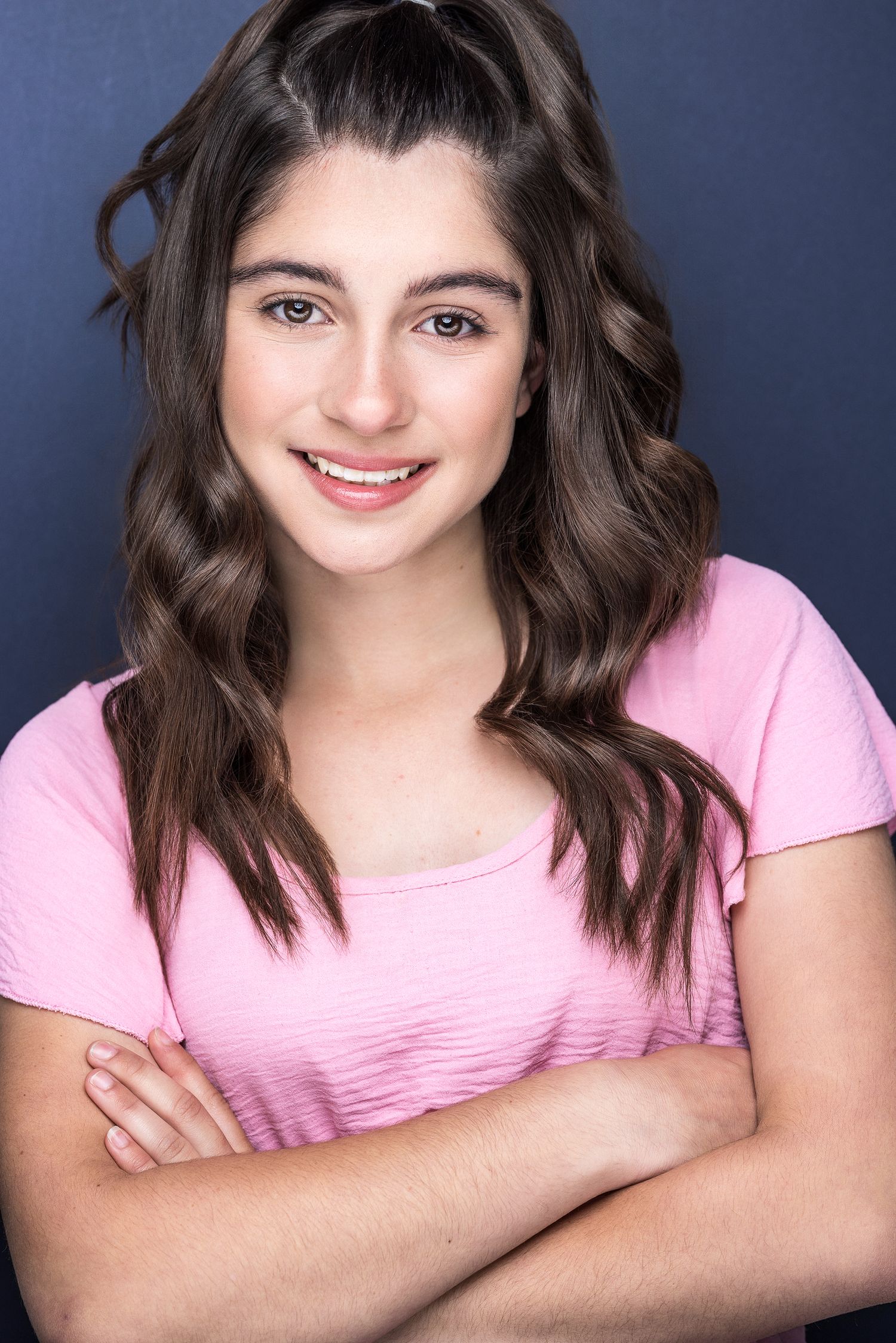 Young woman with brown wavy hair smiling confidently, arms crossed, wearing a pink t-shirt against a dark background.