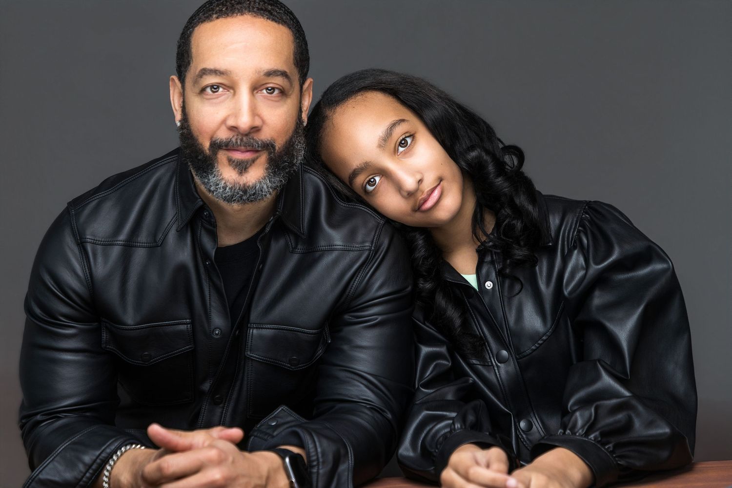 A man and young woman in matching black leather jackets pose together for a professional portrait on gray background.
