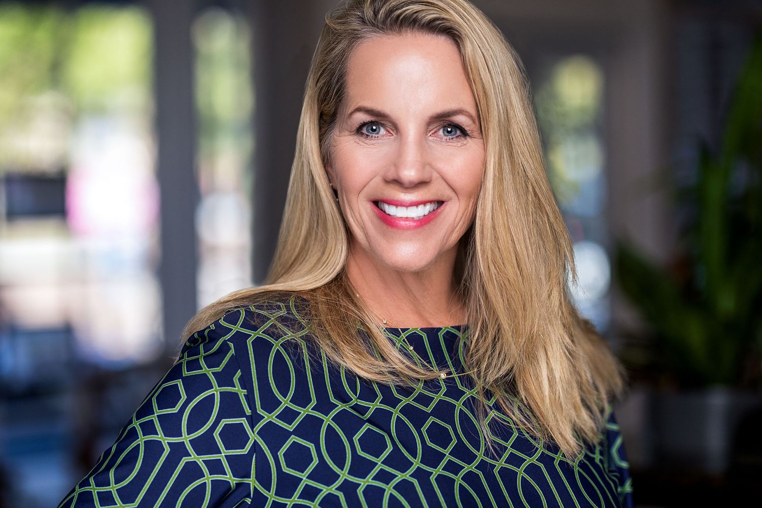 Smiling blonde woman in a green and navy patterned top poses for a professional headshot indoors.