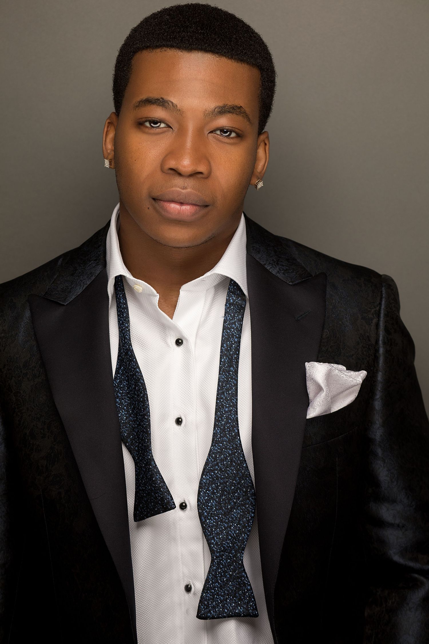 Young Black man in black tuxedo jacket with bow tie and pocket square poses for formal portrait.