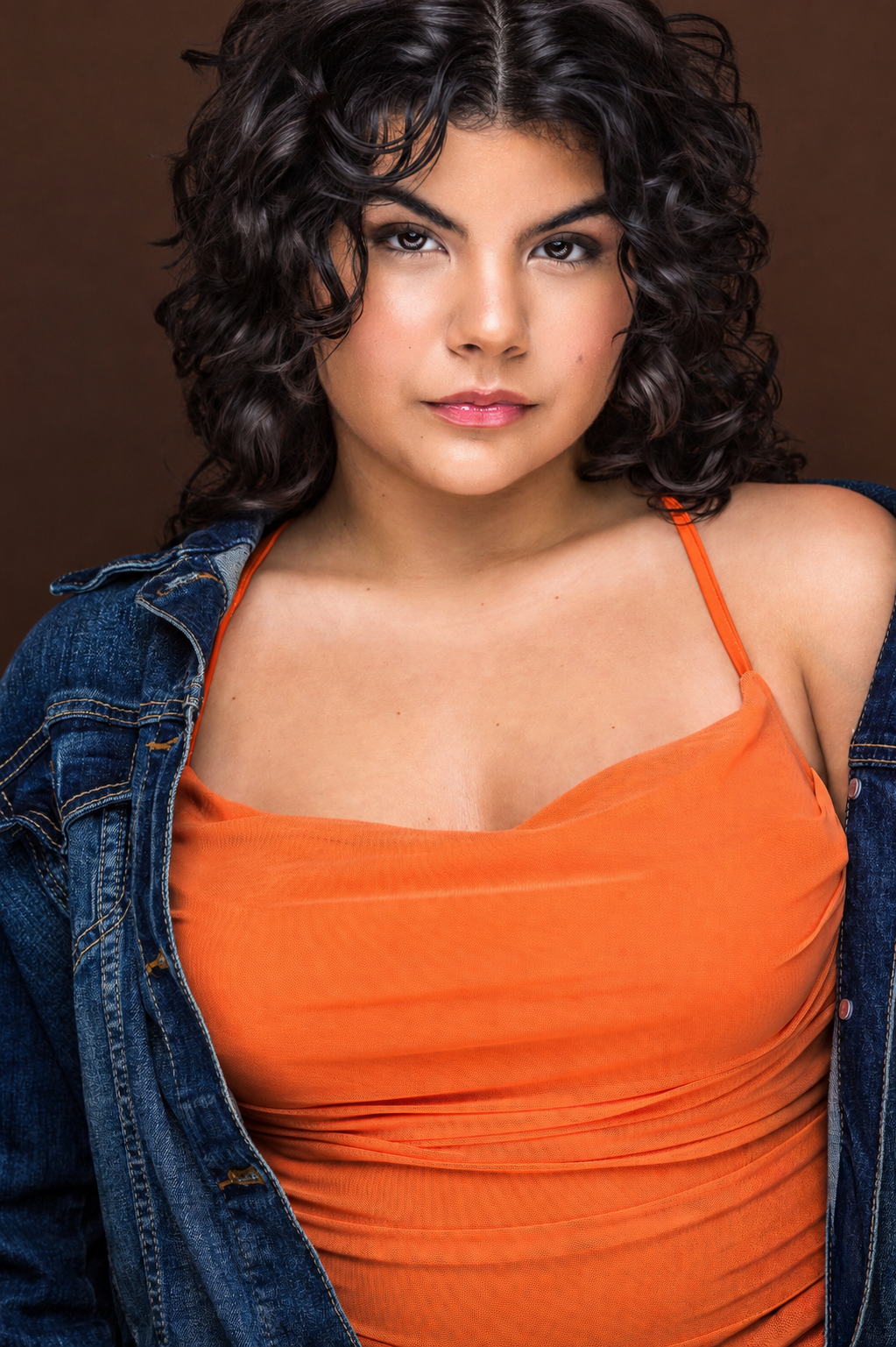 Young woman with dark curly hair in an orange top and denim jacket poses for a professional headshot on brown backdrop.