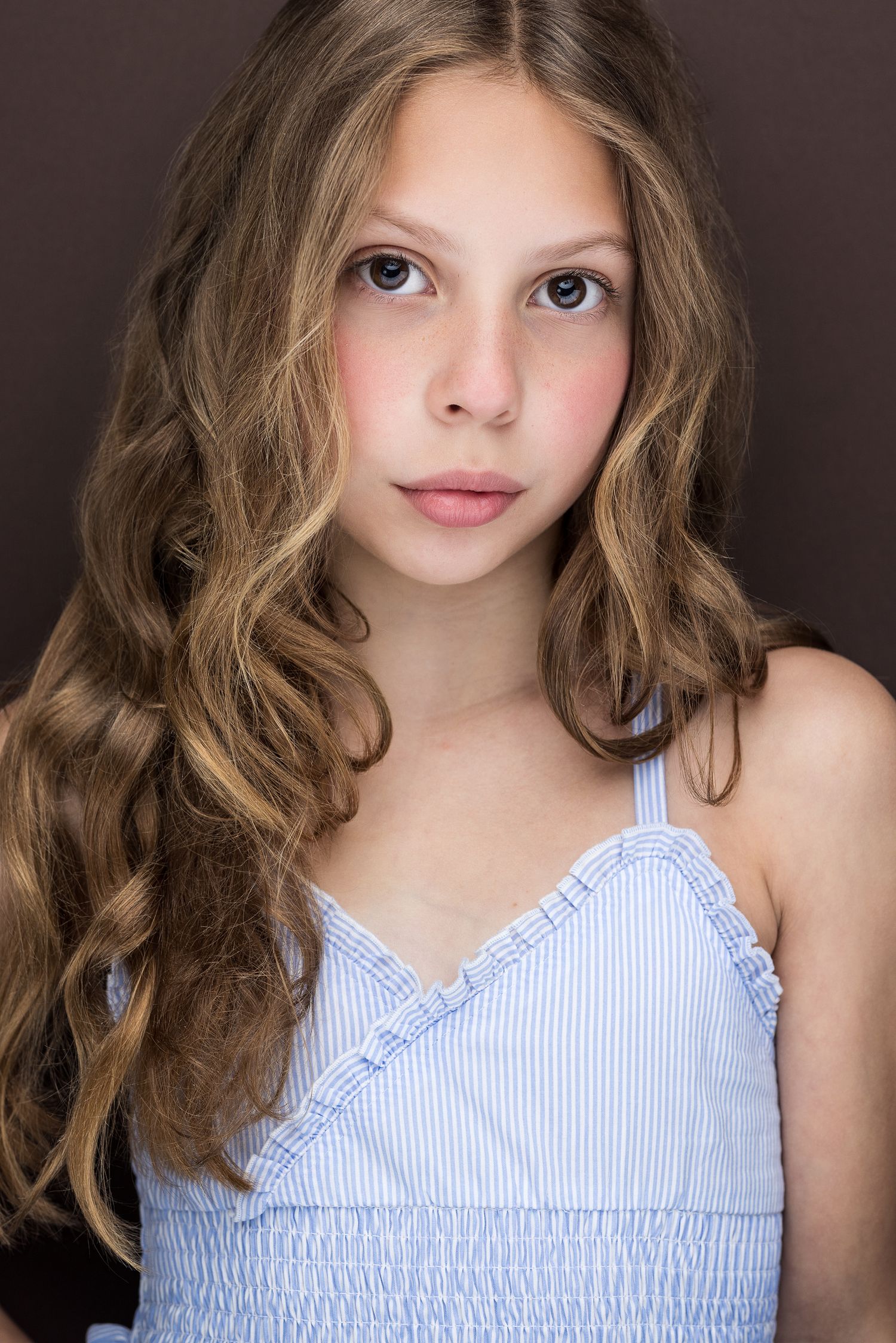 Young girl with long wavy brown hair in light blue sundress against dark background.