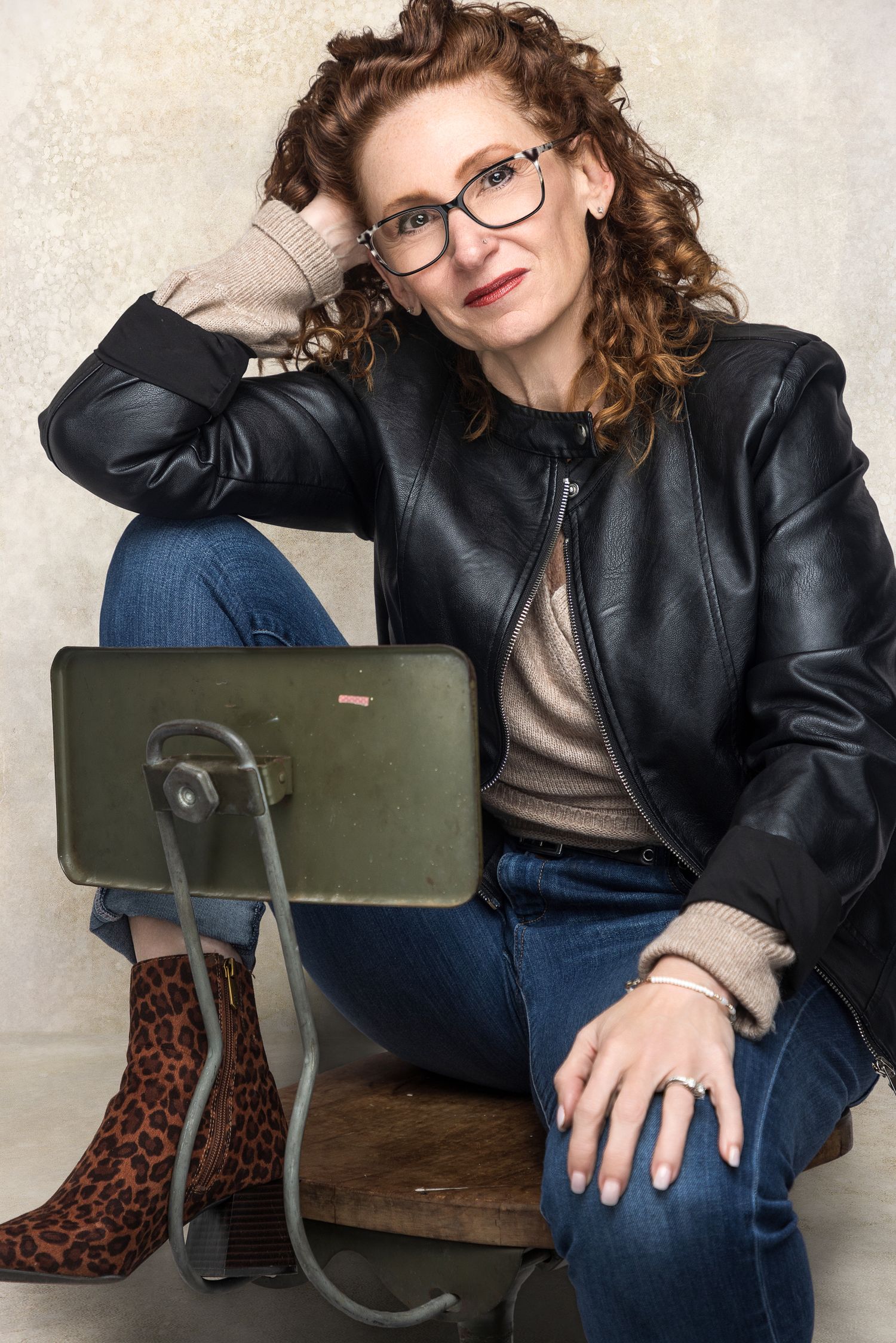 Woman in leather jacket, jeans, and leopard boots sits relaxed with glasses and curly hair beside a vintage tablet stand.