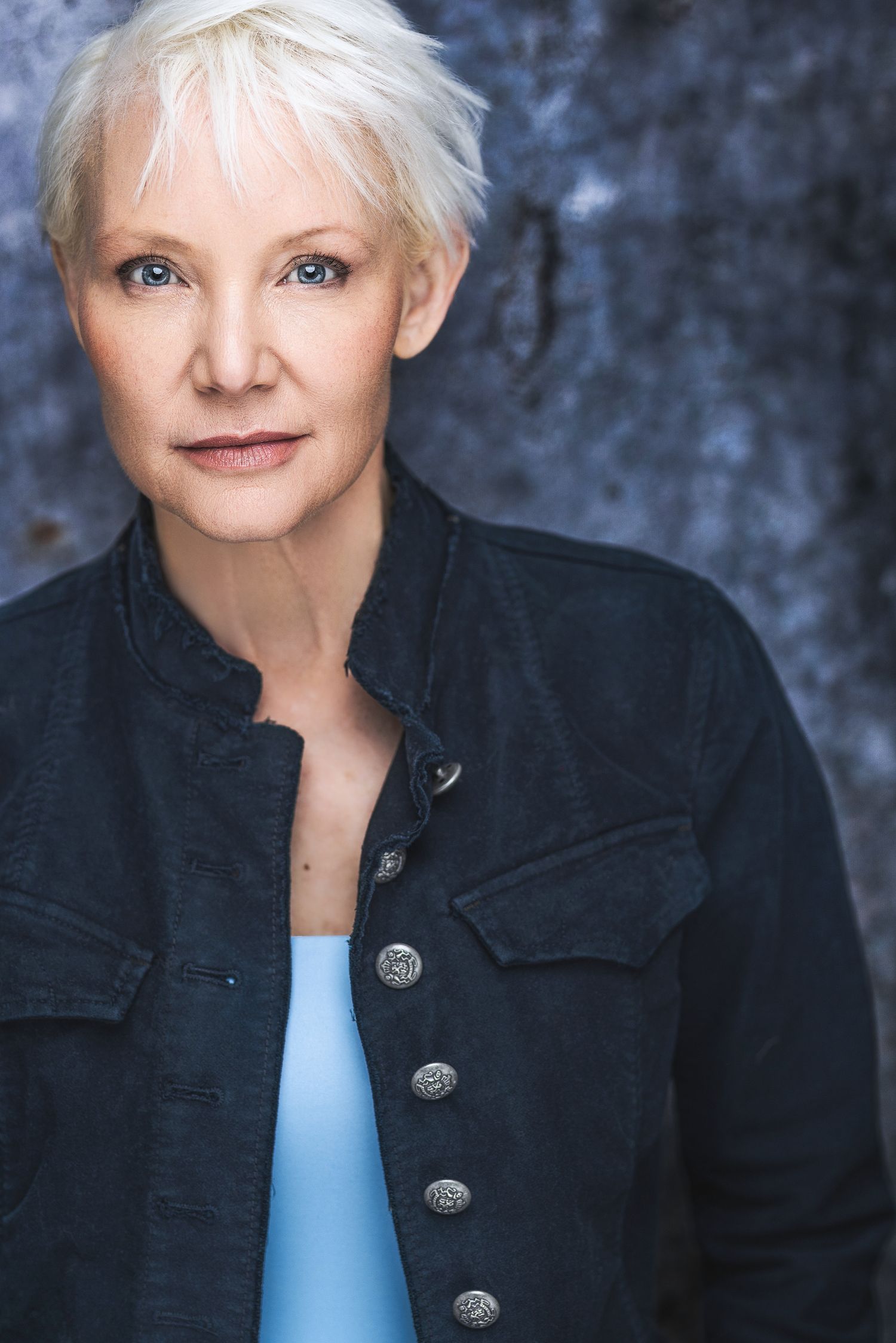 A mature woman with short white hair and blue eyes wearing a black denim jacket over a light blue top.