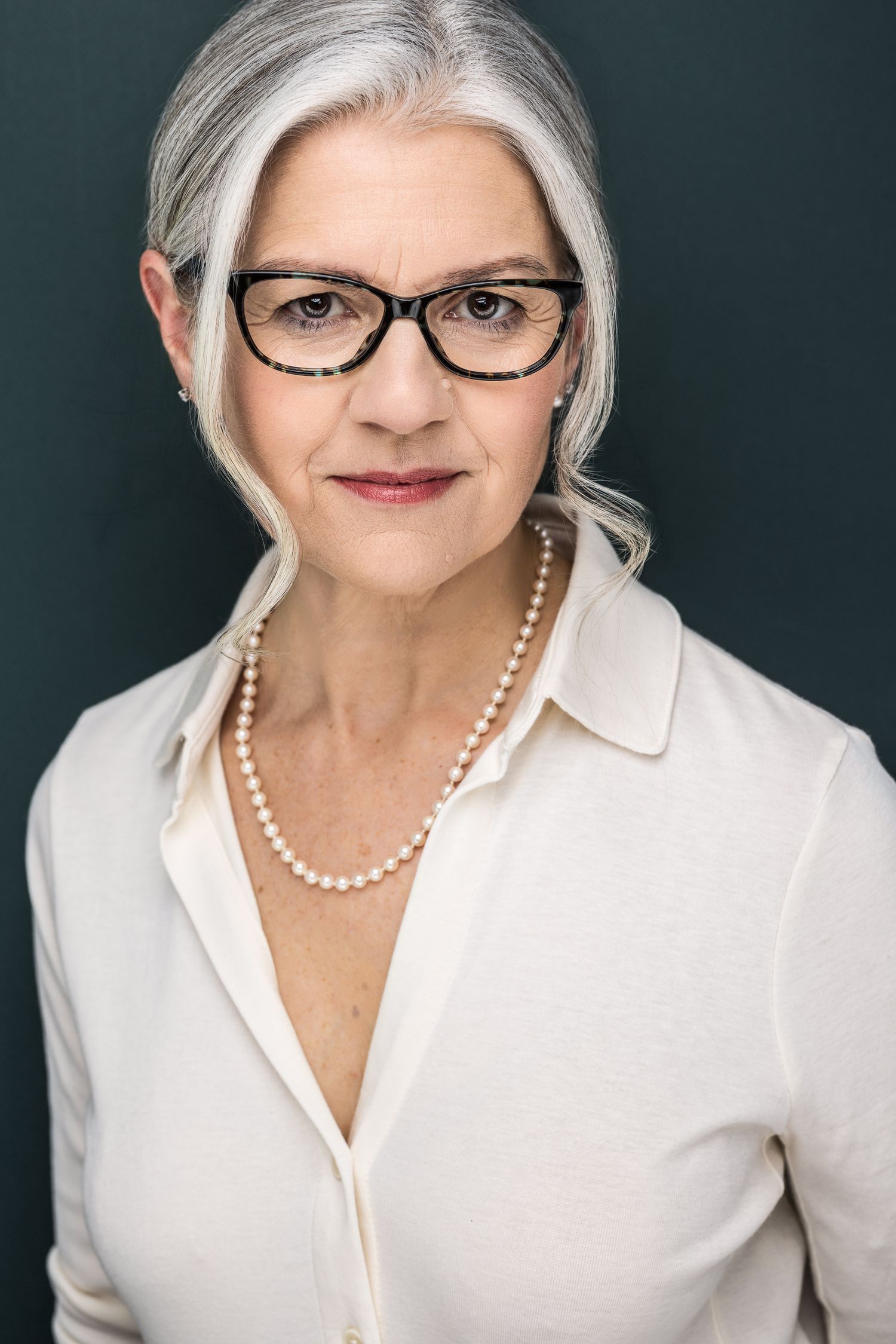 Elegant older woman with gray hair, black glasses, pearl necklace, and white blouse poses for a professional headshot.